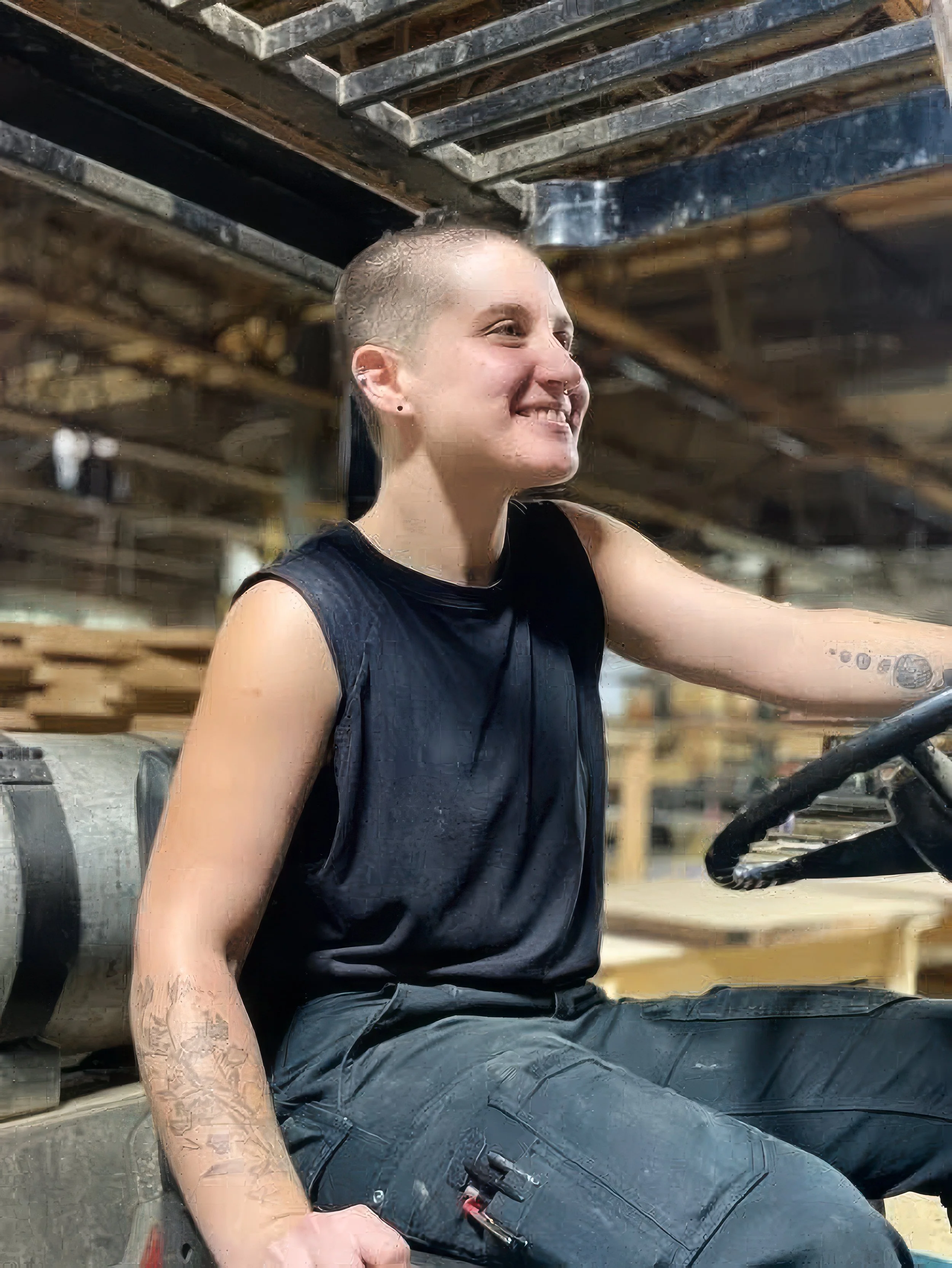 A person with a shaved head sitting on a construction vehicle, smiling and wearing a black sleeveless shirt and black cargo pants, inside an industrial workshop with wooden and metal structures.