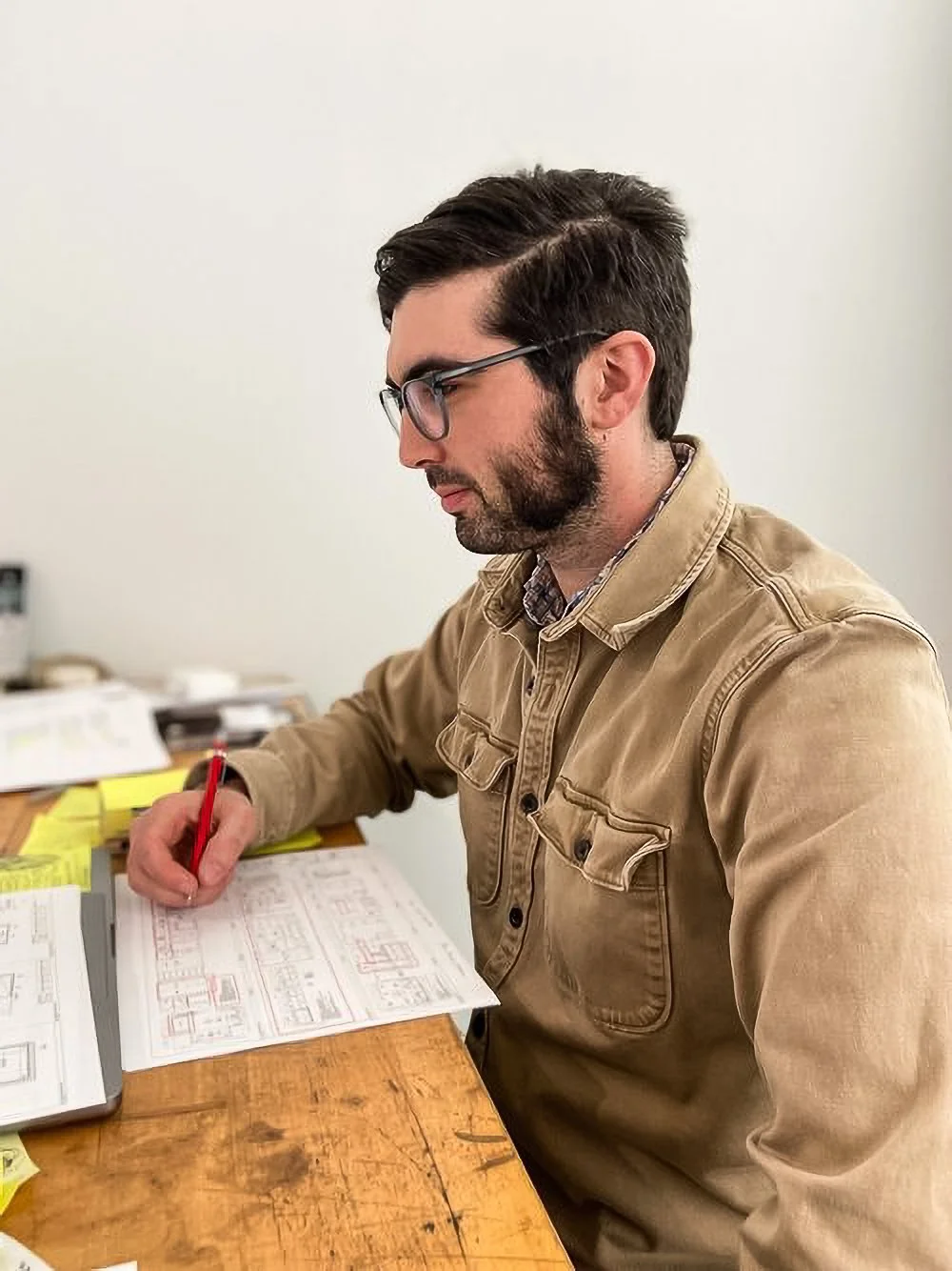 Man wearing glasses and a tan jacket sitting at a wooden desk, working with a pencil and architectural blueprints.