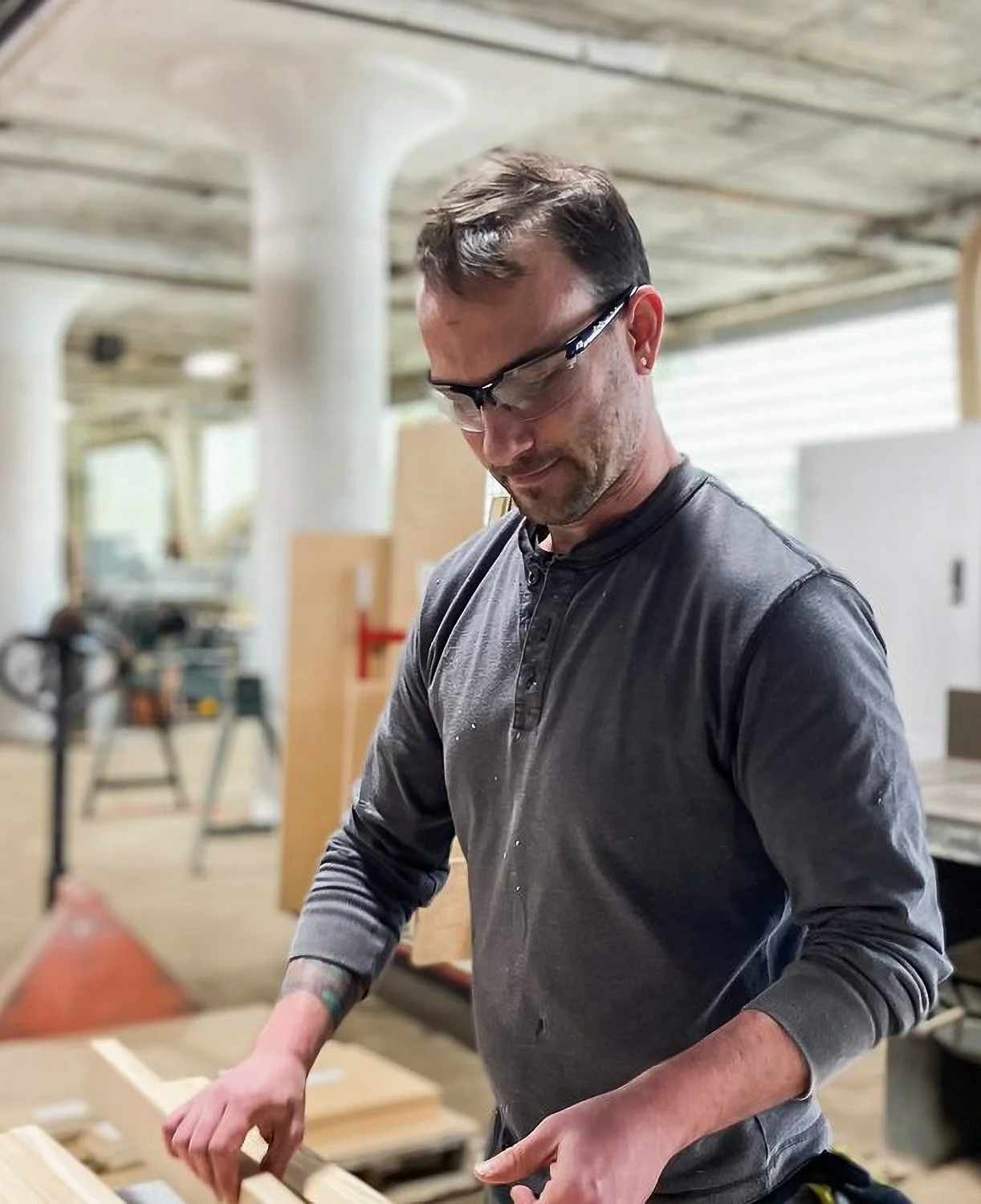 A man wearing safety glasses and a long-sleeved shirt working in a woodworking shop, handling a piece of wood.