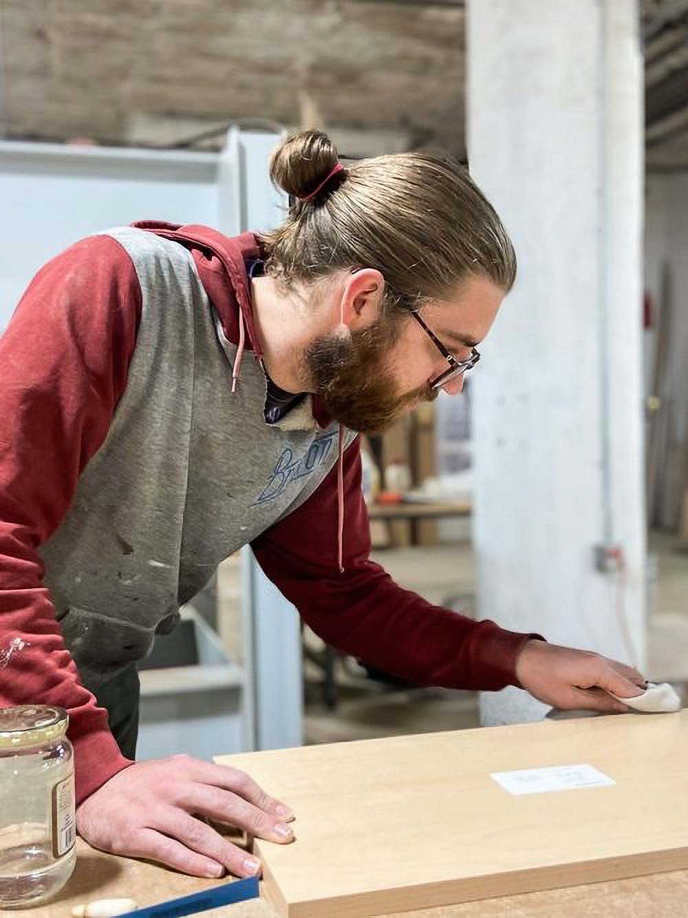 A man with long hair tied back and glasses, wearing a gray and red hoodie, is leaning over a wooden table in a workshop, sanding or polishing a piece of wood with a cloth.