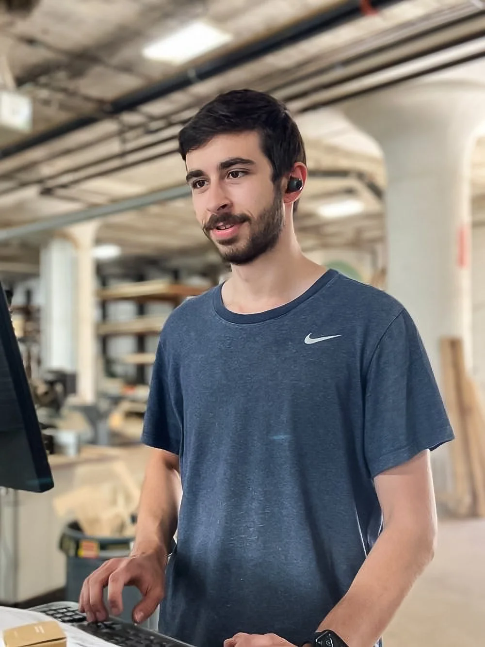 A young man with dark hair and a beard is working at a computer in a woodworking workshop, wearing a dark blue Nike t-shirt and wireless earbuds.