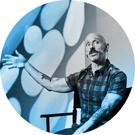 A bald white man with tattoos on his arms is speaking at a panel event, in front of a backdrop of large scale pills of varying sizes. He's gesturing to the left, with a kind but serious look on his face.