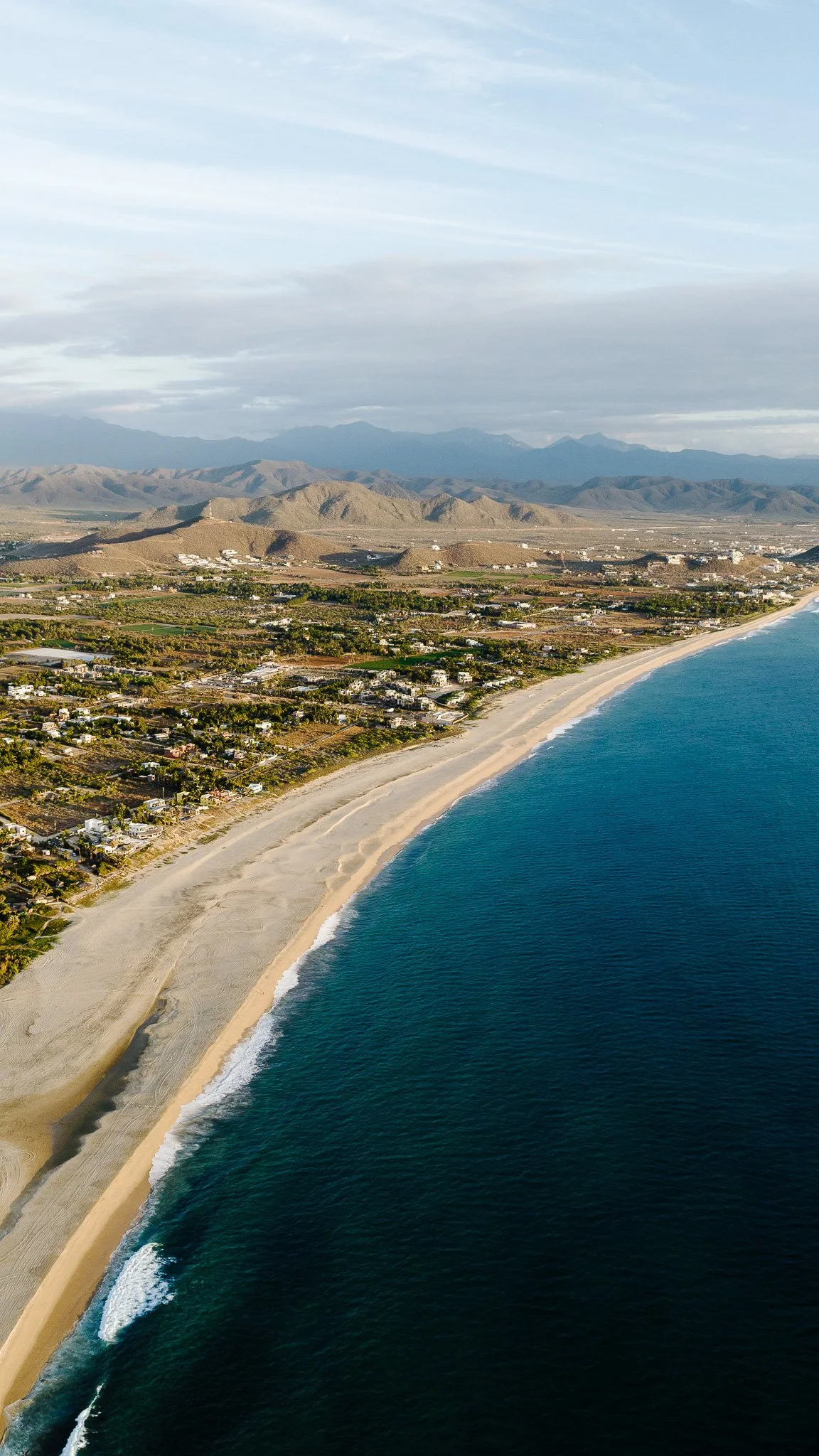 Aerial view of a sandy beach along the coastline with waves crashing, residential area behind the beach, hilly terrain further inland, and mountains in the background under cloudy skies.