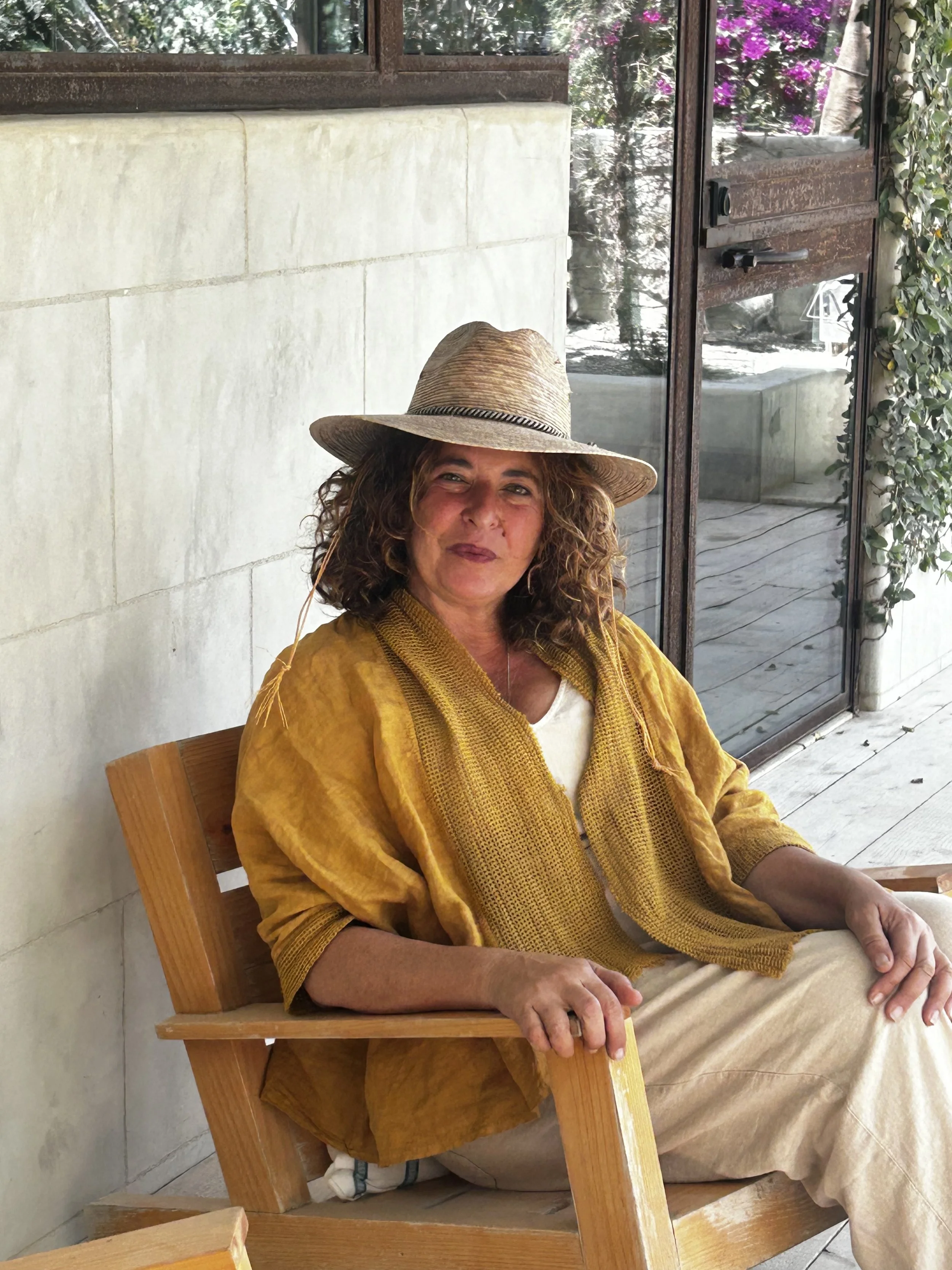 A woman with curly hair wearing a wide-brimmed straw hat and a yellow shawl, sitting on a wooden chair near a glass door with greenery and purple flowers outside.
