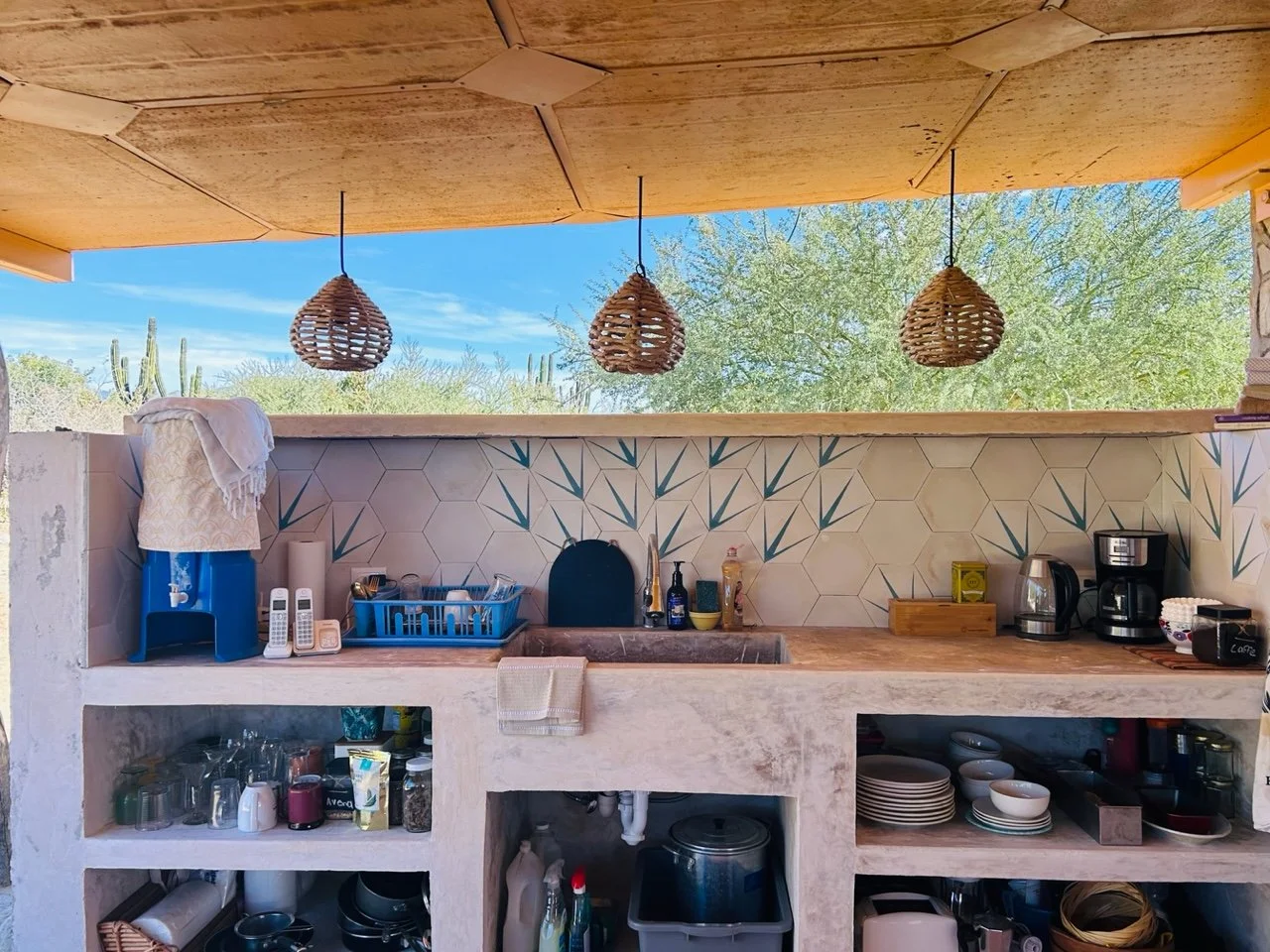 Outdoor kitchen with a hexagon-tiled backsplash, wooden countertop, and woven pendant lights, featuring a sink, dish rack, coffee maker, electric kettle, and shelves with dishes and glasses, with desert landscape and cacti visible in the background.