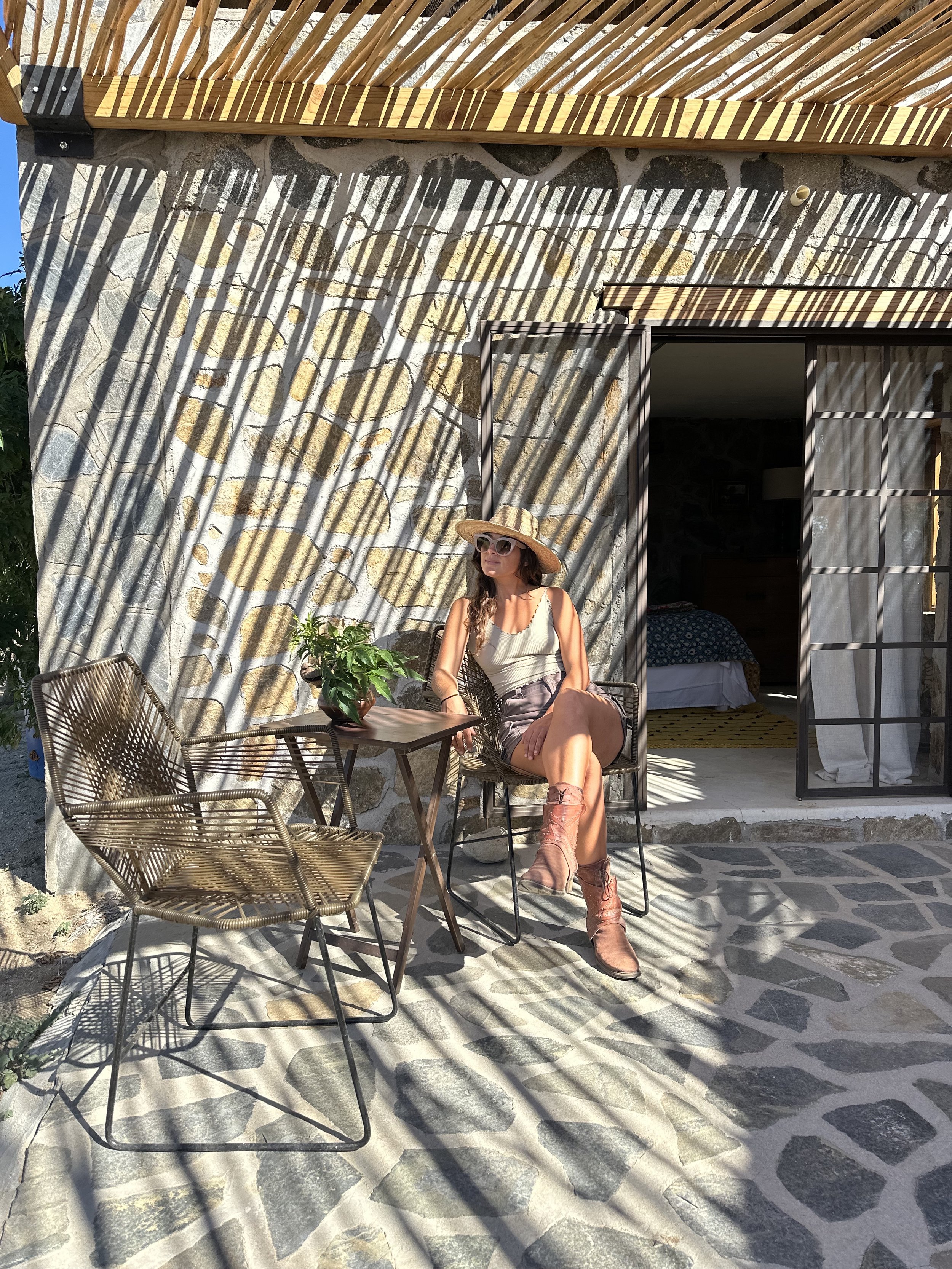 A woman sitting on a chair on a patio with stone flooring, wearing a wide-brimmed hat, sunglasses, a sleeveless top, shorts, and boots. There are two metal chairs and a small table with a potted plant. The patio is next to a building with a stone wal