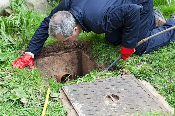 Man examining a sewer pipe in the ground near a manhole cover.