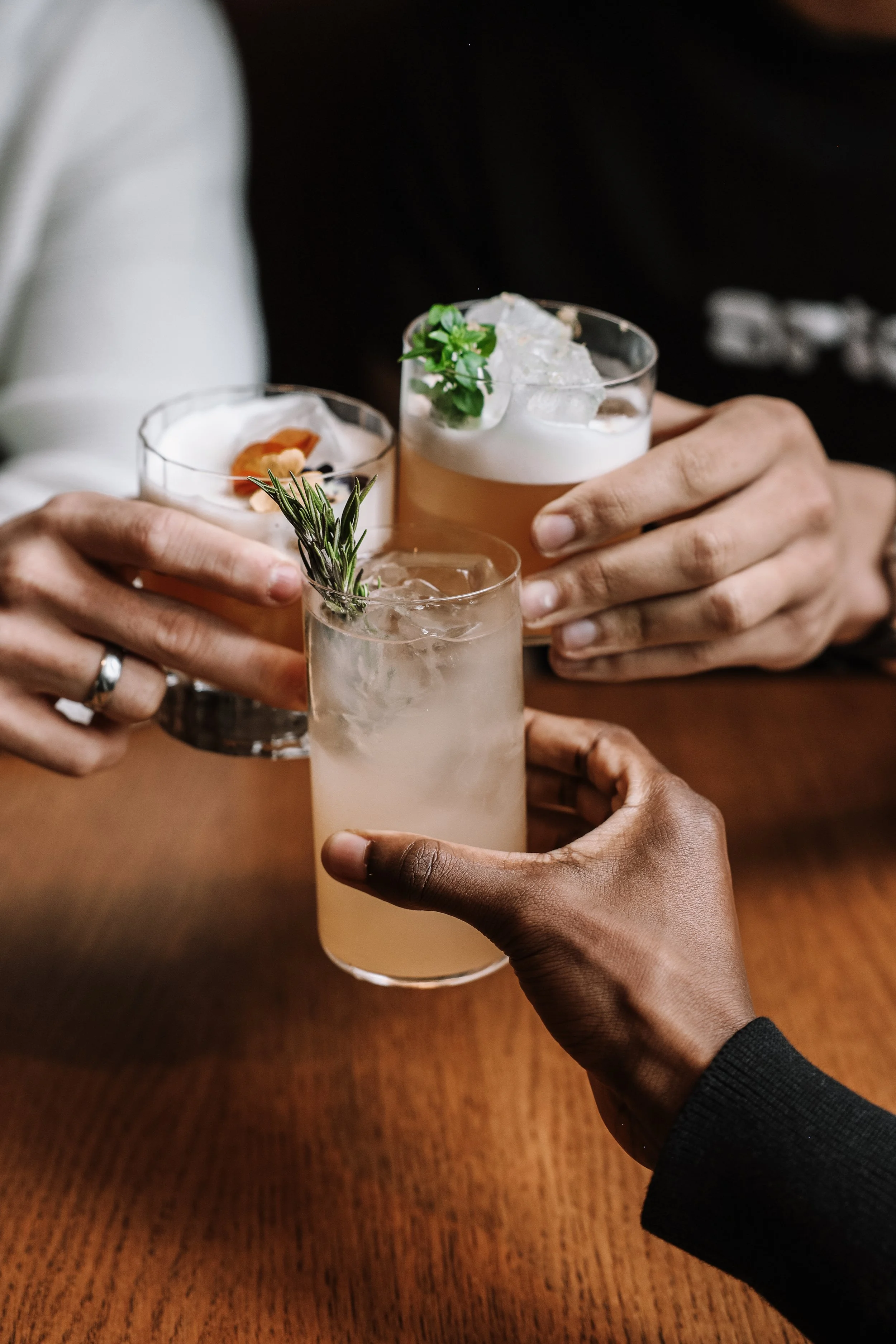 Three people clinking glasses with cocktails on a wooden table, featuring garnishes like rosemary and ice cubes.
