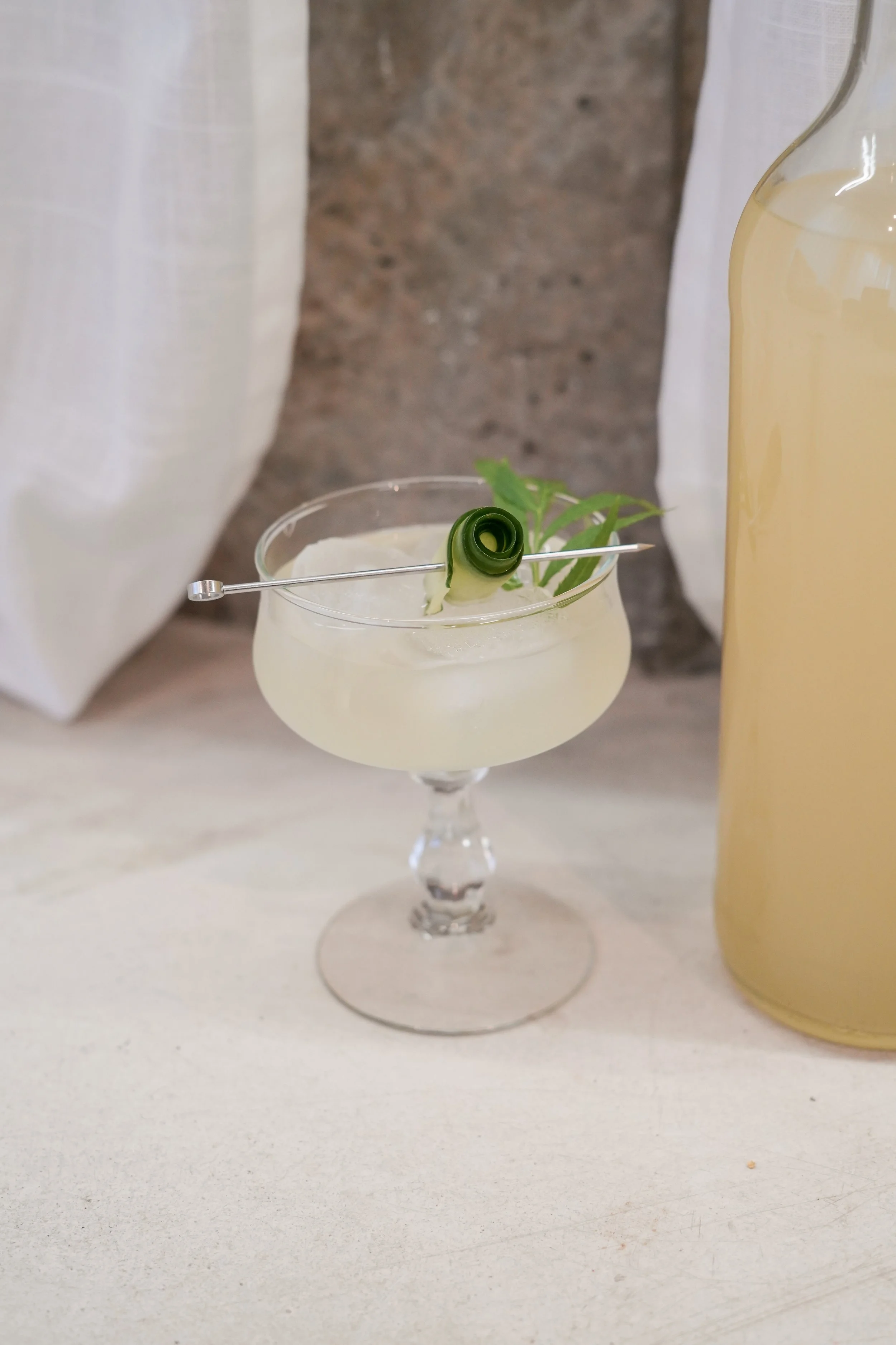 A cocktail with ice in a stemmed glass, garnished with a cucumber ribbon and a sprig of green herbs, next to a bottle of light-colored liquid on a white surface.