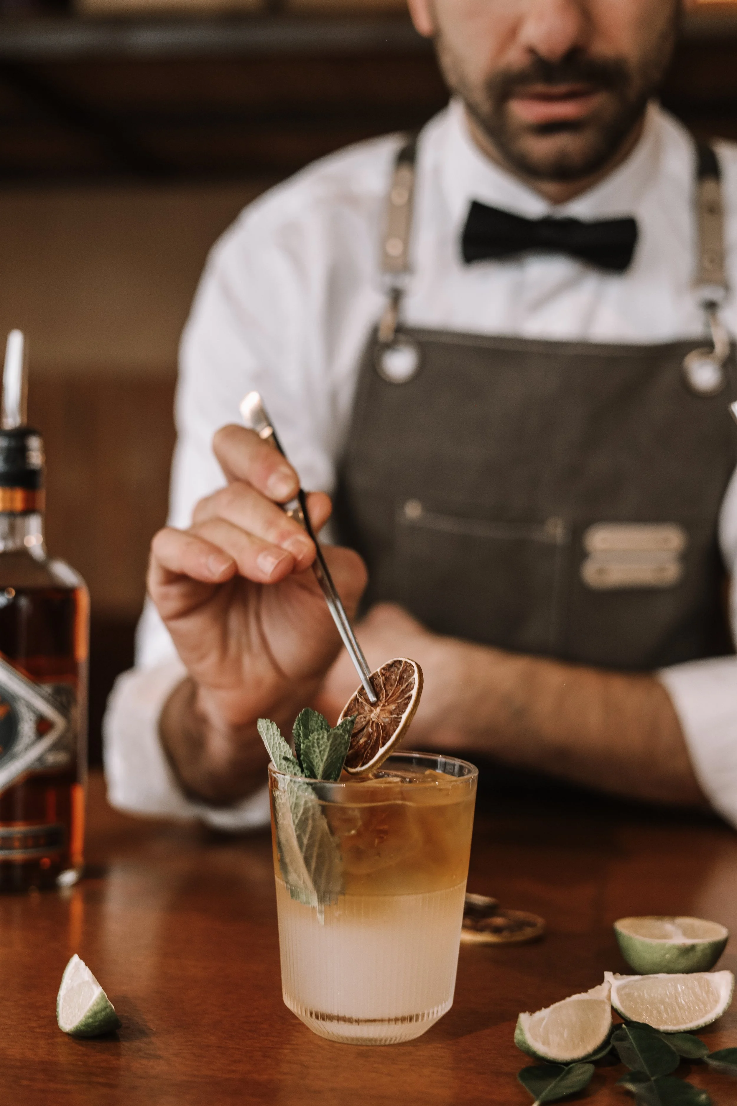 Bartender garnishing a cocktail with a dried citrus slice and mint leaves, with lime wedges on the table.