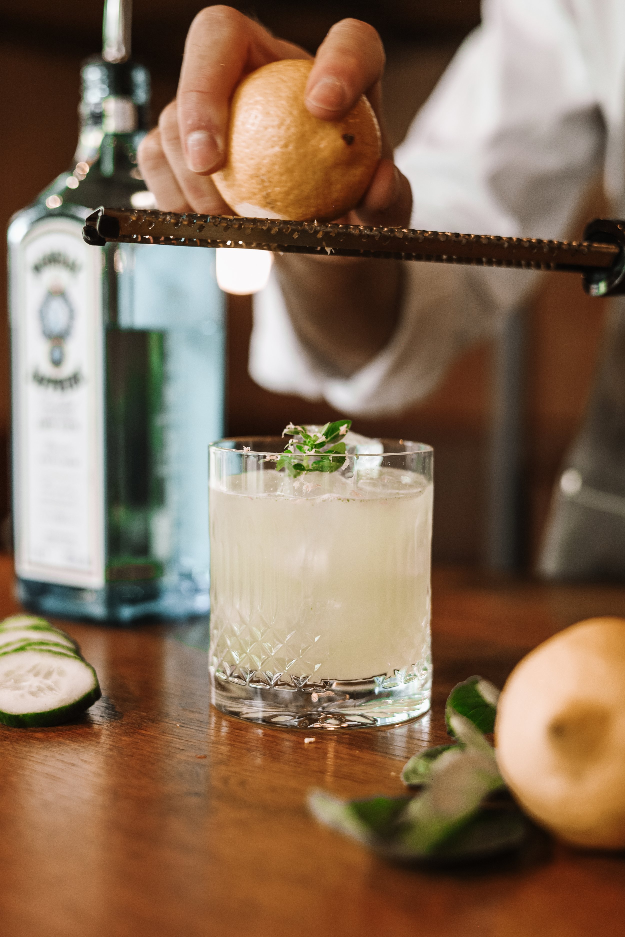 Close-up of a hand zesting a lemon over a cocktail in a glass, with a bottle in the background and cucumber slices on a wooden table.
