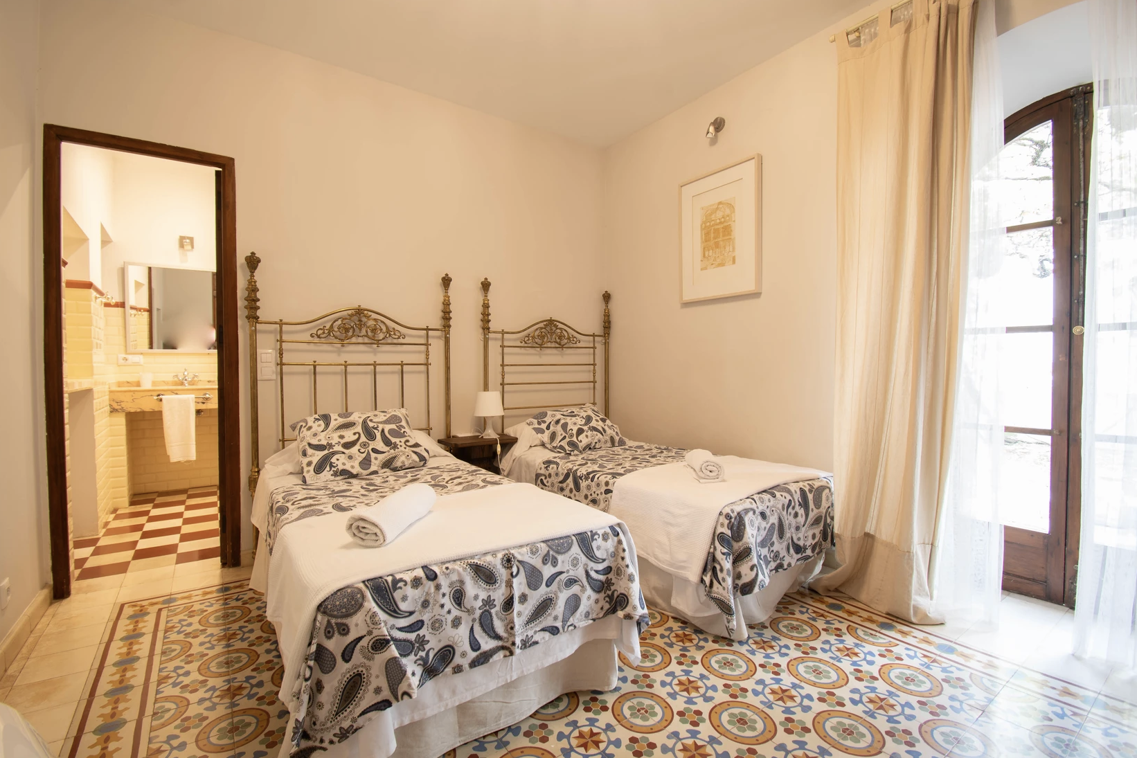 Bright and elegantly styled twin bedroom at Casería de Milán, featuring vintage decor, tiled flooring, and a cozy en-suite bathroom, offering a comfortable stay during the retreat in Granada, Spain.