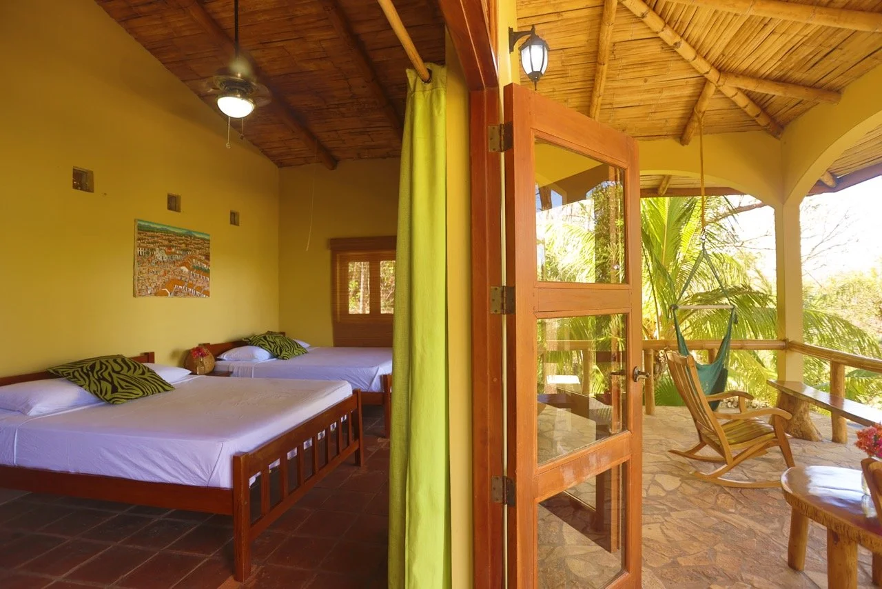 Interior of a bright guest room at Villa Laguna with queen beds, wooden ceiling, and patio doors opening to a private outdoor seating area – yoga retreat accommodation in Nicaragua
