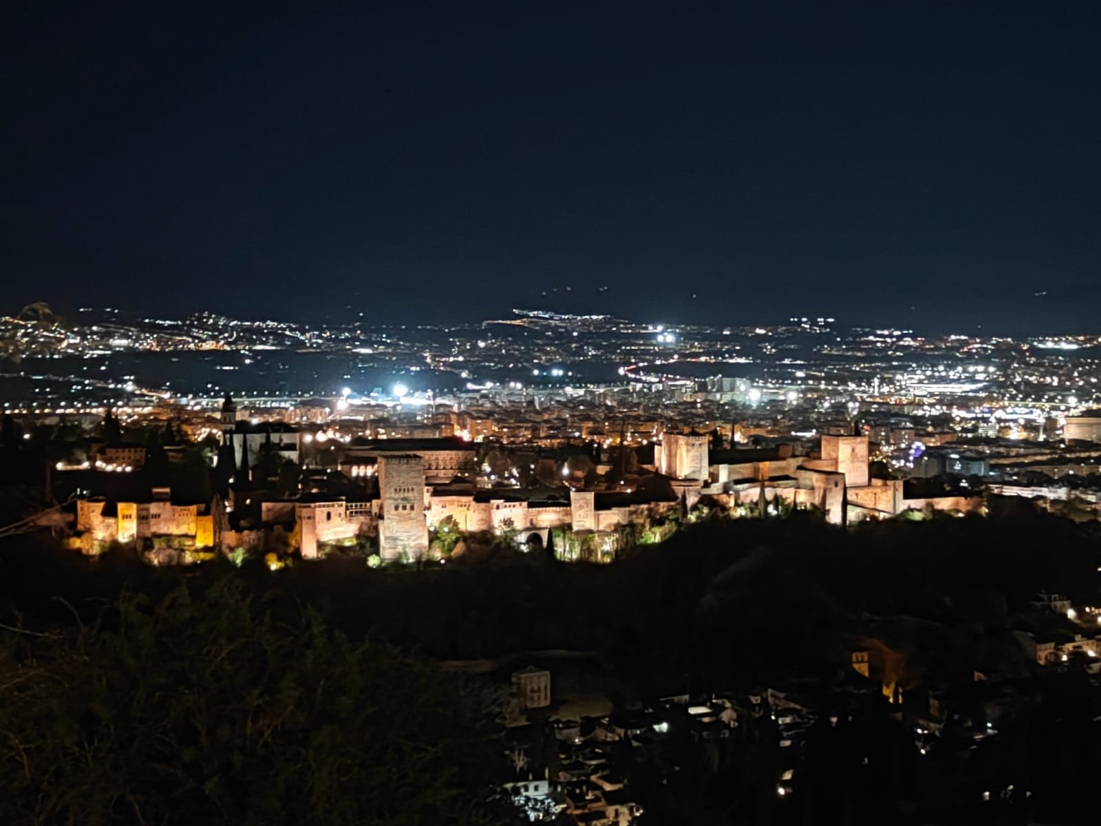 The Alhambra Palace beautifully illuminated at night, overlooking the city of Granada, Spain, with sparkling city lights in the background, showcasing the palace’s majestic presence after dark.