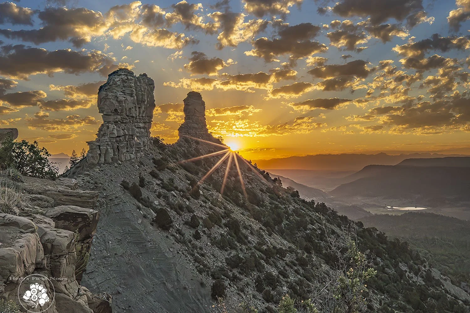 Sunset over rugged mountain landscape with two large rock formations and rainbows in the background.