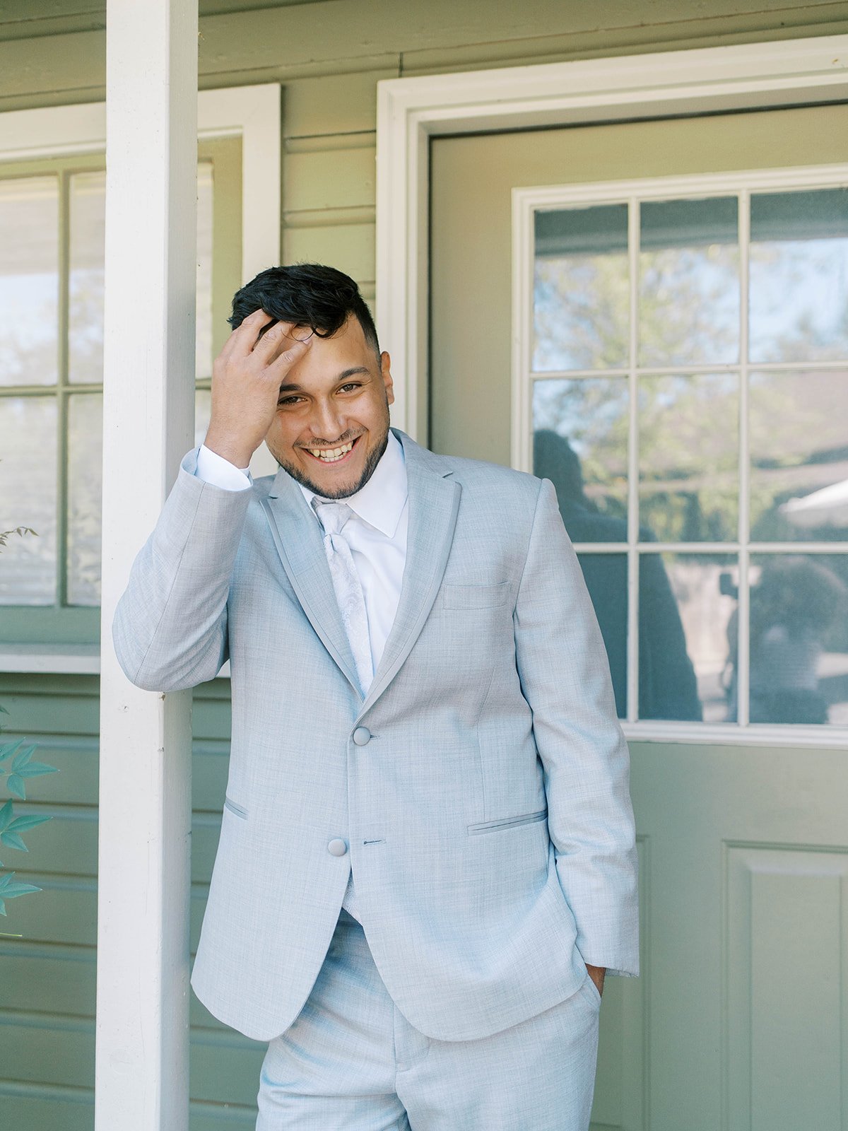 Smiling groom in light gray suit standing on porch before wedding ceremony.