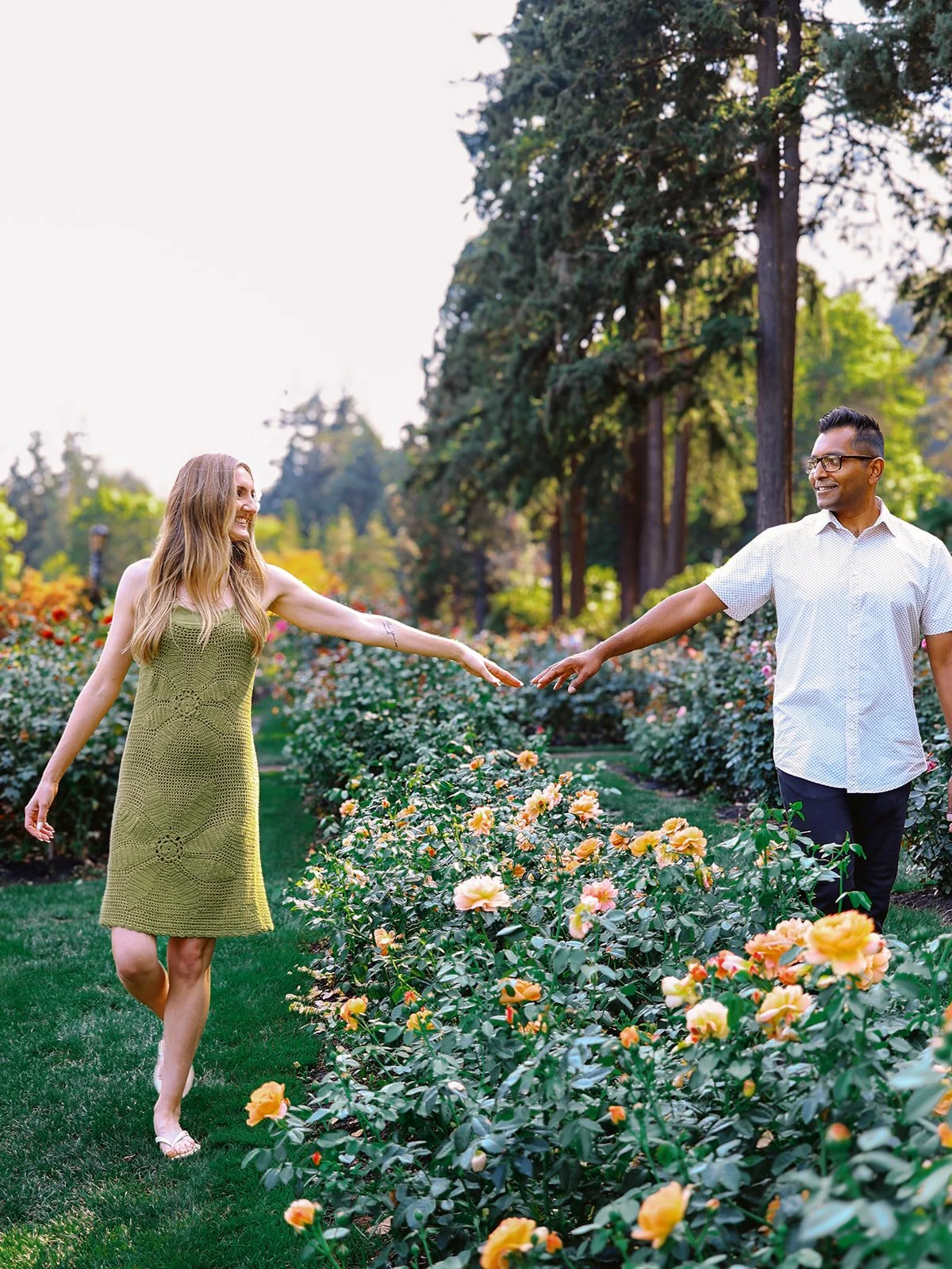 A couple reaching toward each other across a row of peach roses in a lush garden, smiling as their fingertips nearly touch.