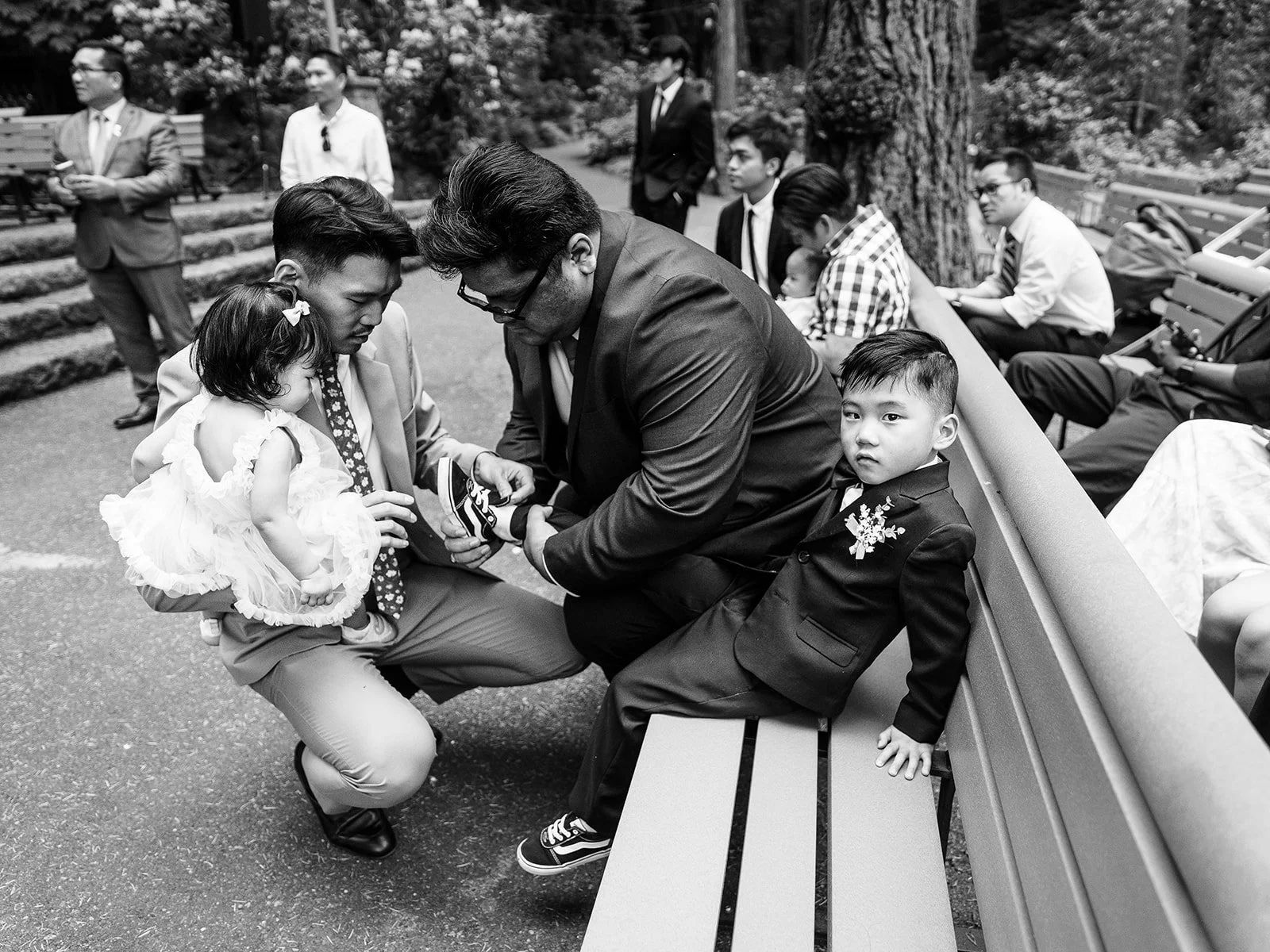 Black and white candid of two men crouching to tend to the ring bearer's shoe on a bench, one also holding a toddler flower girl, with wedding guests milling in the background at The Grotto