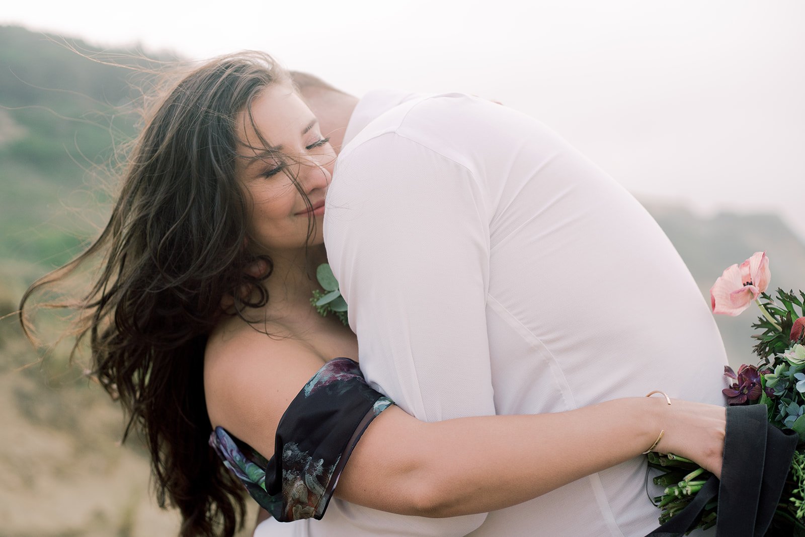 Woman embracing partner on Oregon Coast cliff, bouquet visible, wind-swept hair and soft light