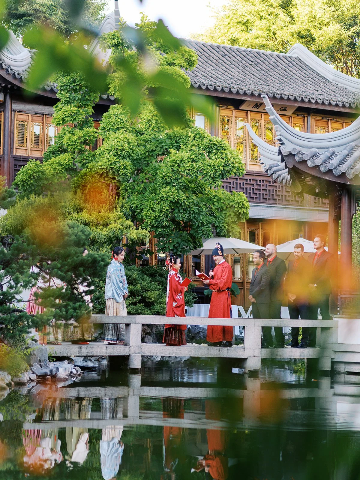 The groom in red robes reads from a book during the ceremony on the bridge, the bride beside him and groomsmen looking on, the garden and pavilion glowing warmly behind them.