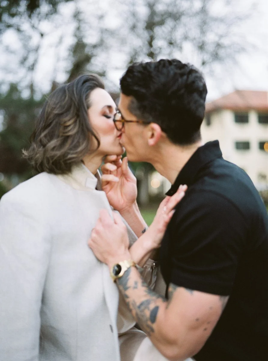 Intimate close-up of couple kissing, woman in cream coat, man in black shirt with tattoos and gold watch