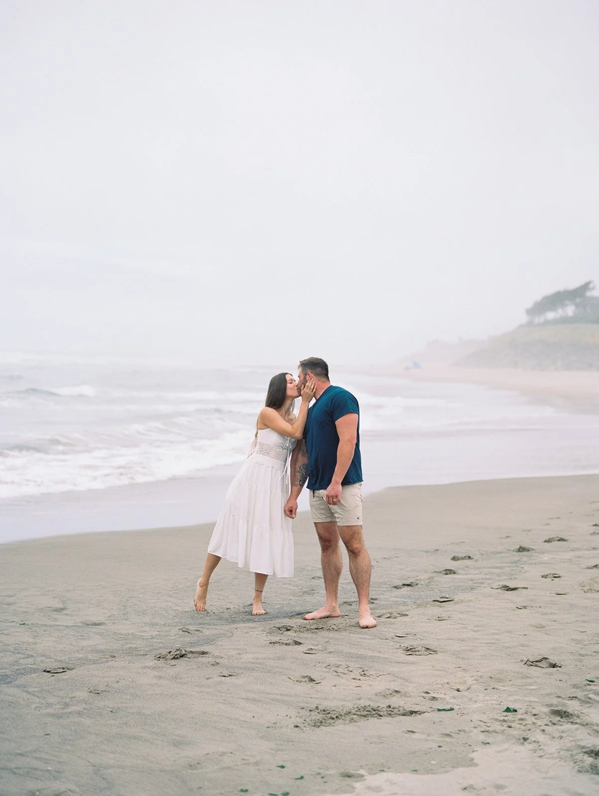 Woman in white dress kissing man on misty shoreline with waves rolling in behind them.