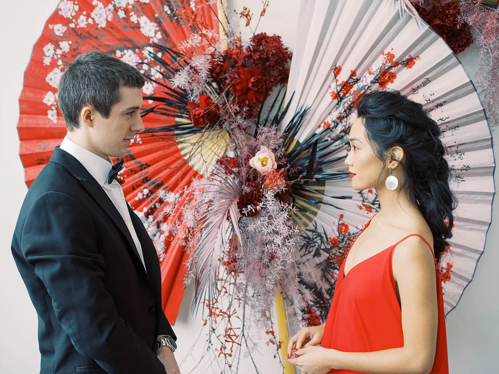 Couple in formal wedding attire face one another before red and white Asian parasols and florals.