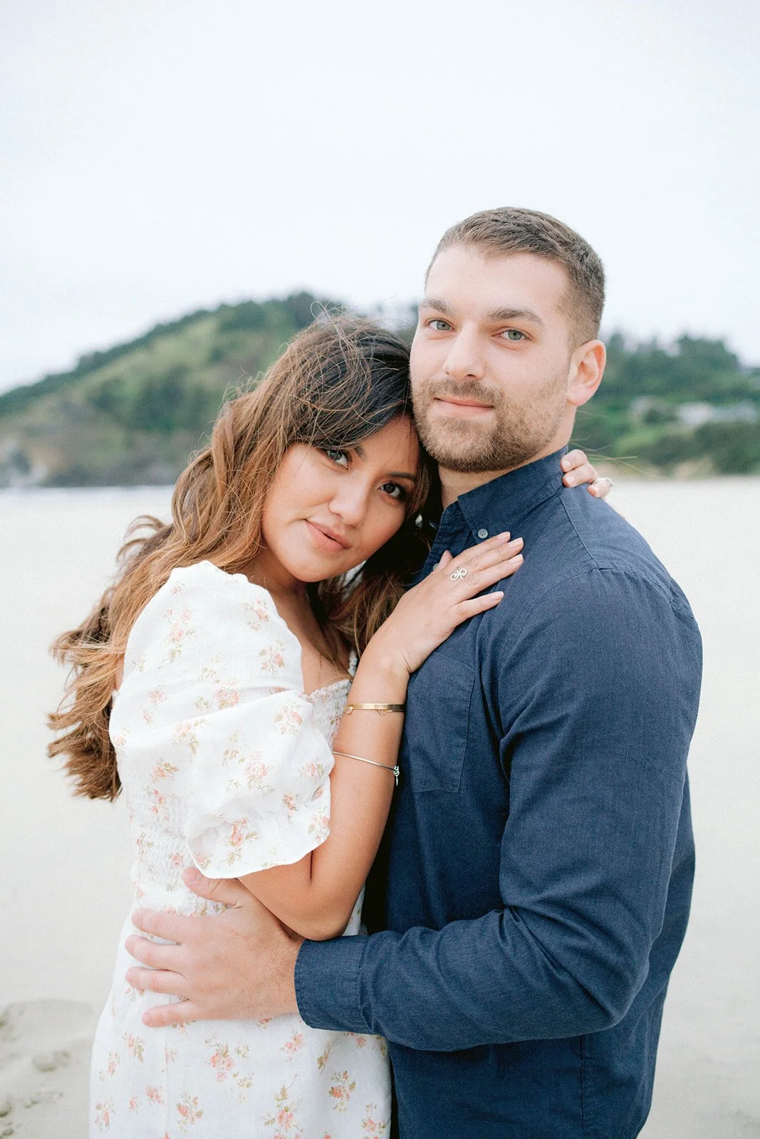 Couple embracing on beach, woman resting head on man’s chest with soft coastal background