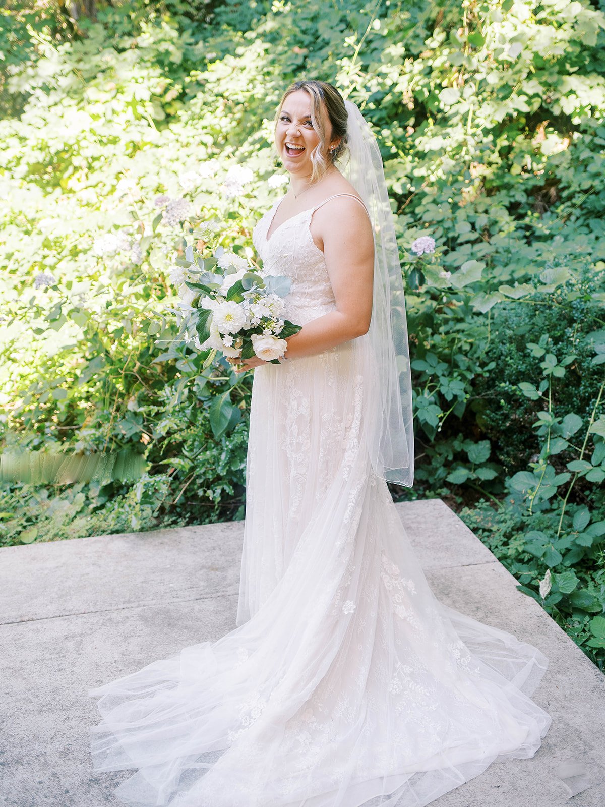 Bride in lace wedding gown holding white garden bouquet outdoors at Abigail’s Garden.