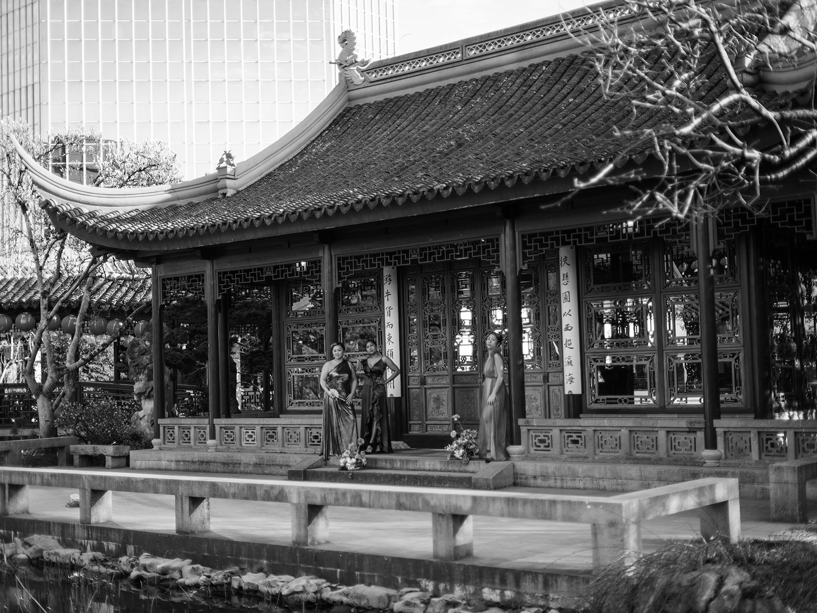 Black and white photo of three women in couture gowns standing on a pavilion overlooking the koi pond at Lan Su Chinese Garden.