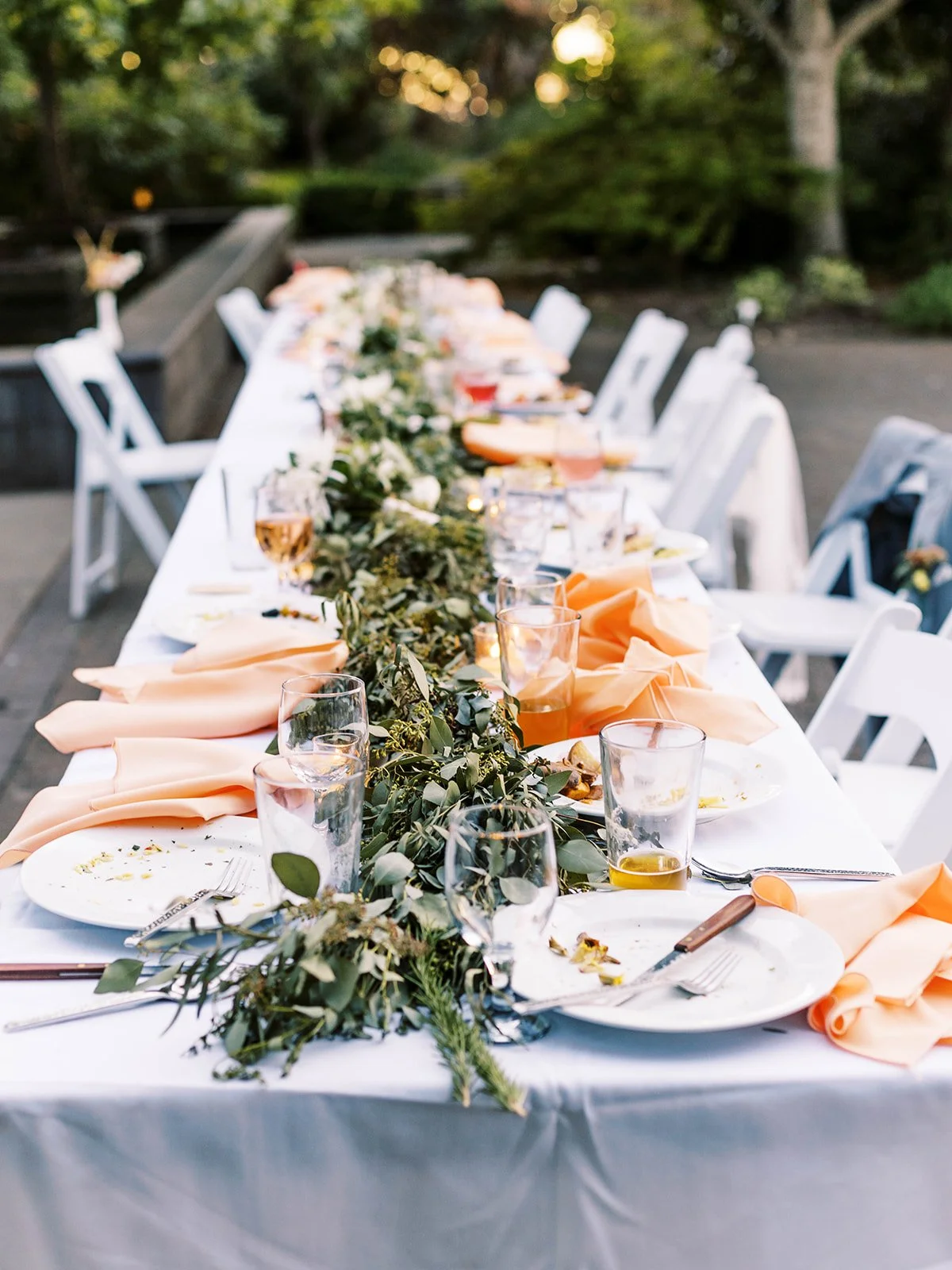 Long reception table with peach napkins, greenery, glassware and white folding chairs after dinner.