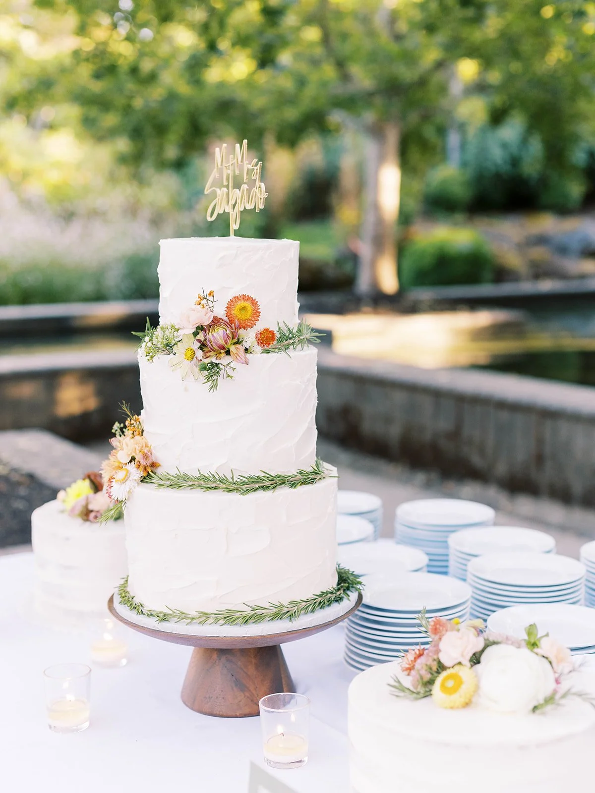 Three-tier white wedding cake decorated with small flowers, greenery and a personalized cake topper.