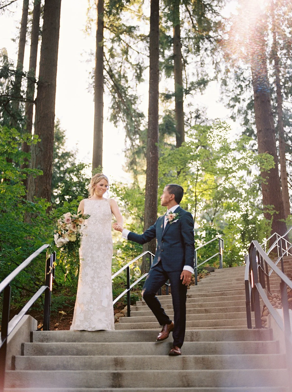 Bride and groom walk hand in hand down stone steps beneath tall trees.