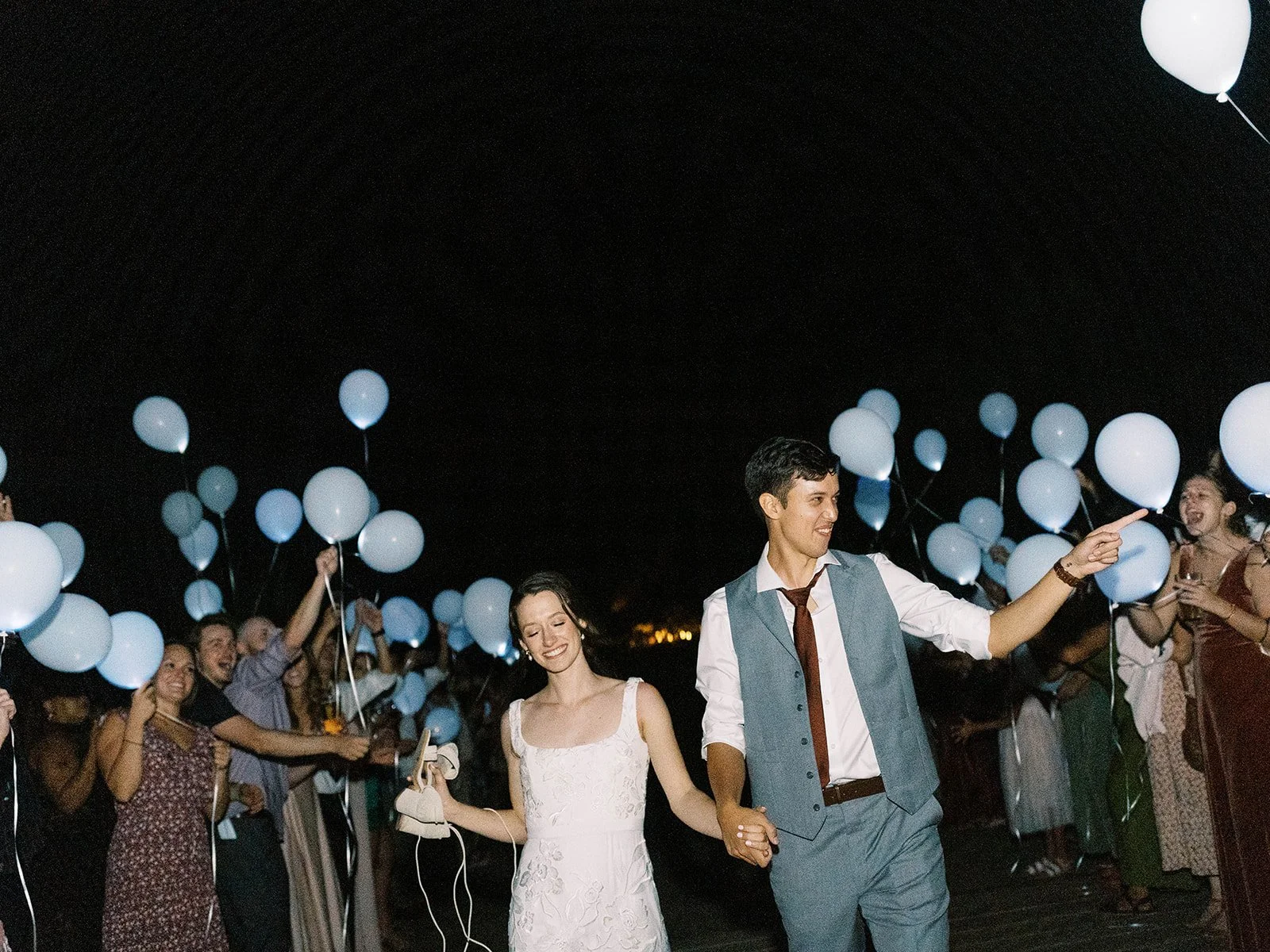 Bride and groom walking hand in hand through a nighttime balloon send-off surrounded by wedding guests.