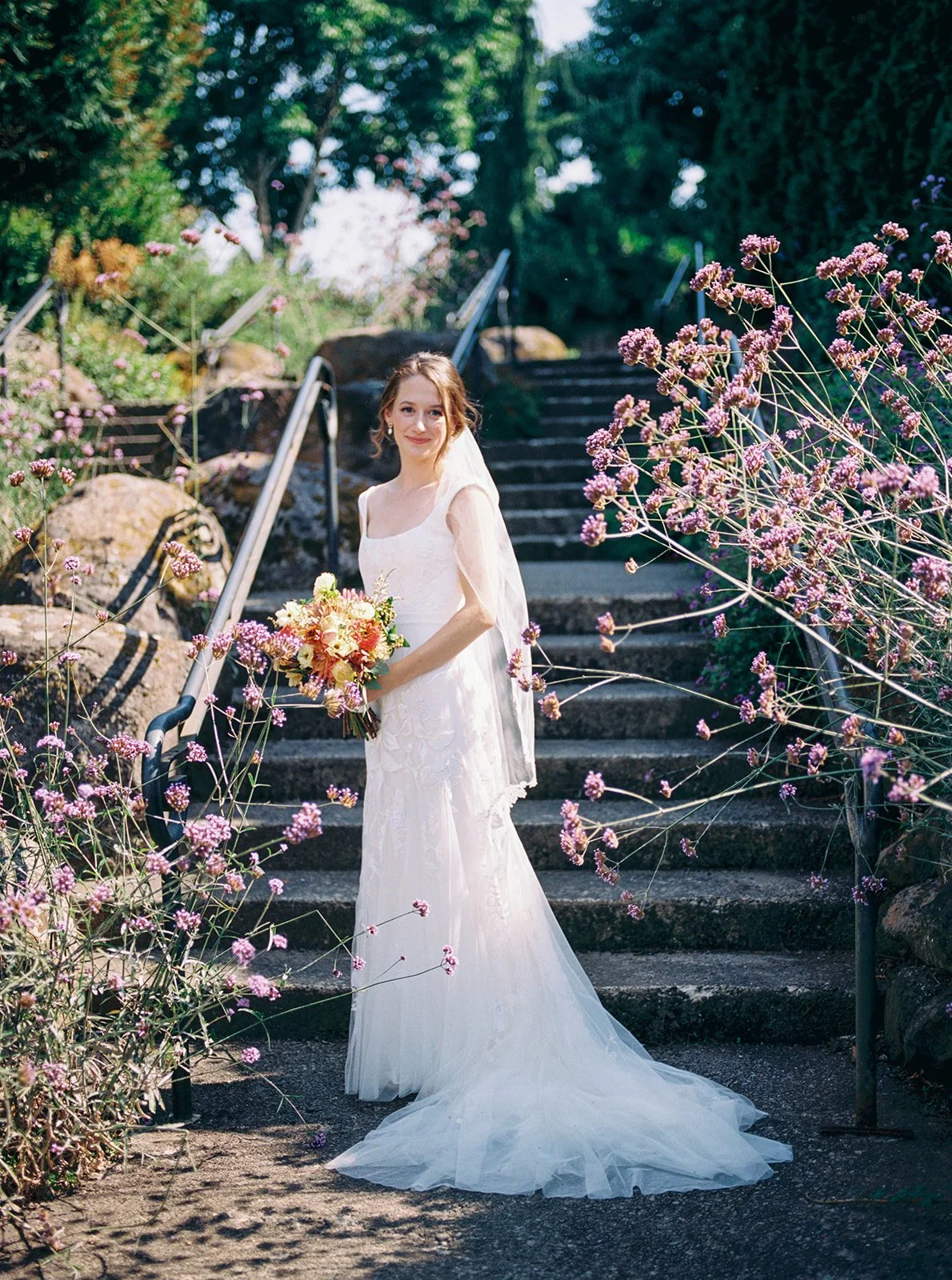 Bride standing on stone steps surrounded by flowers and greenery at Oregon Gardens, holding a bouquet