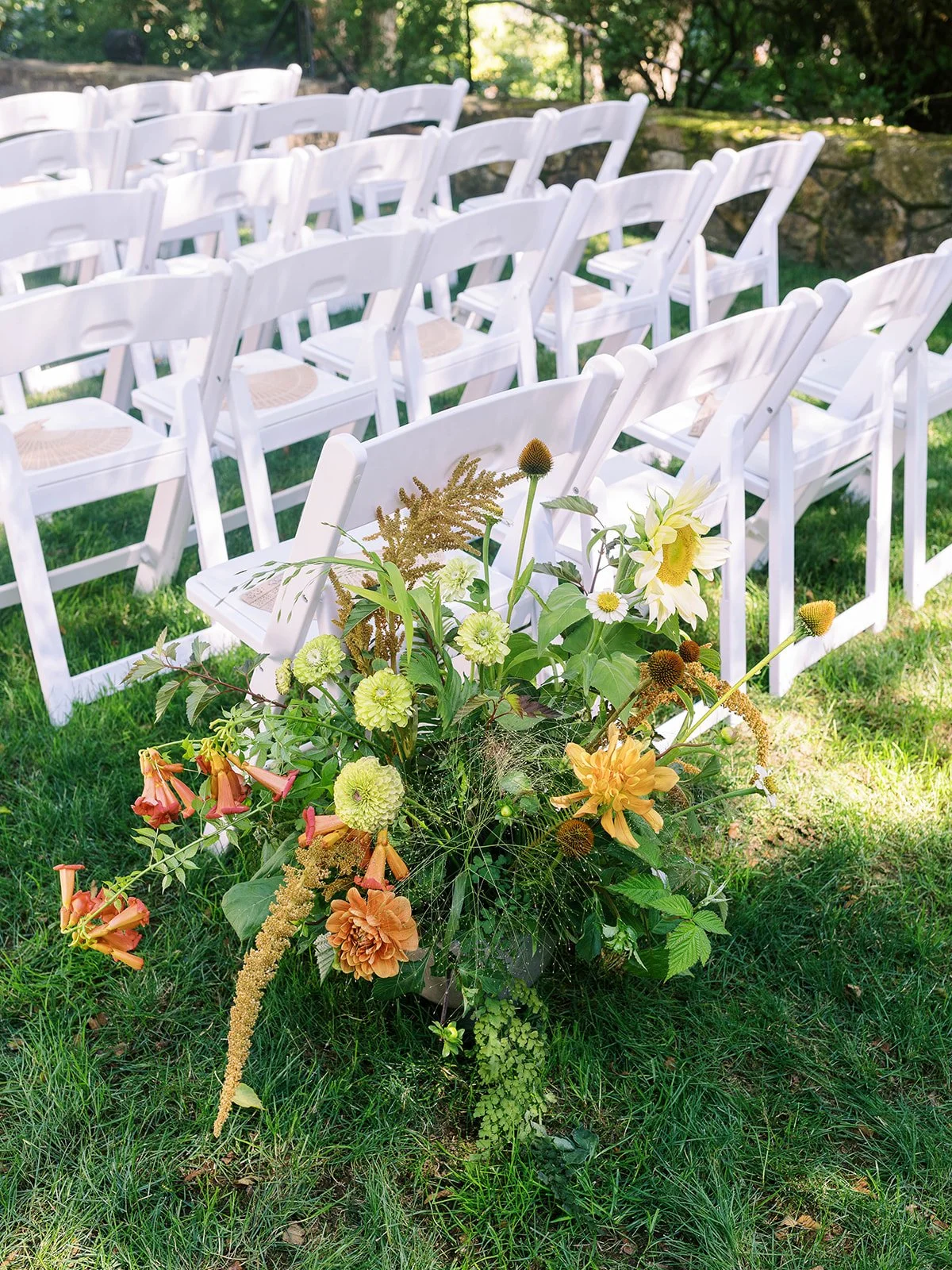 White ceremony chairs beside a low floral arrangement of orange, yellow and green flowers.
