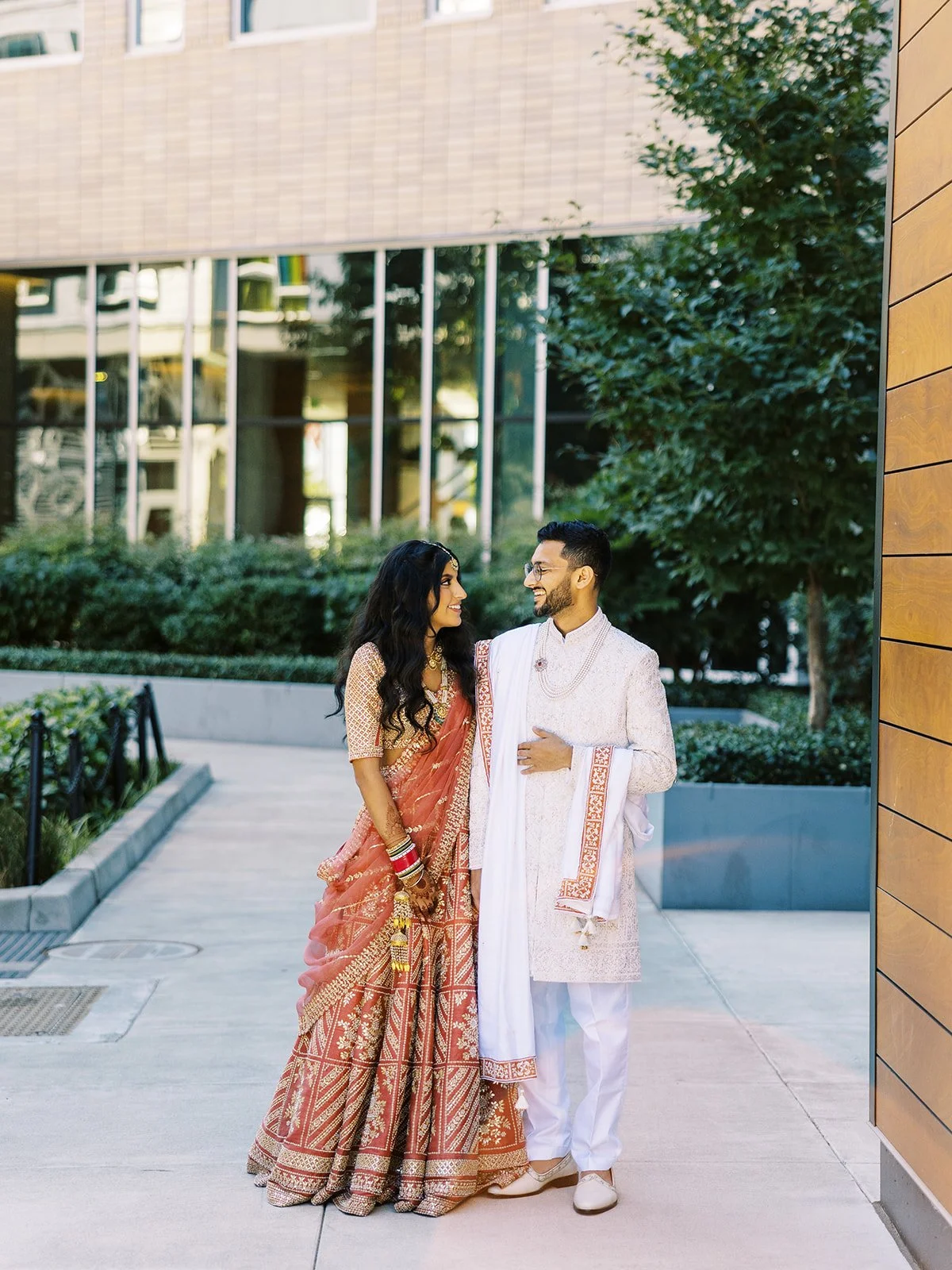 Bride in red and gold lehenga standing with groom in ivory sherwani outside modern wedding venue.