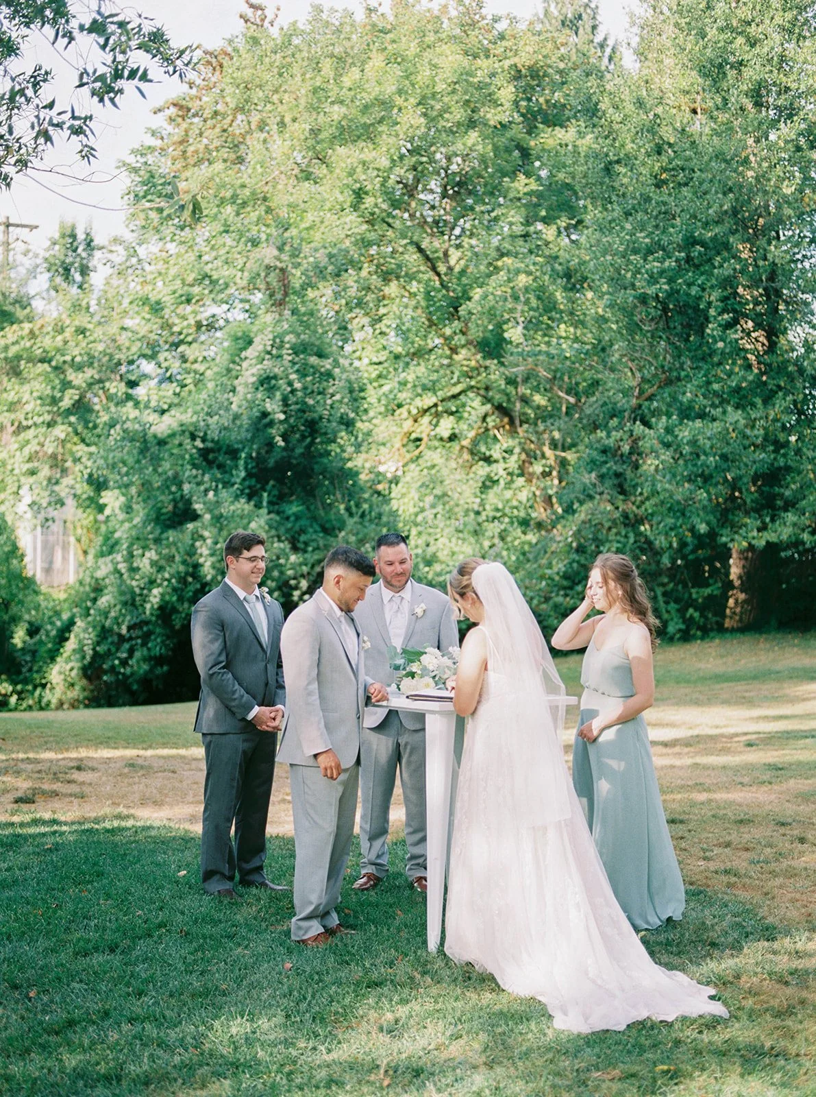 Bride and groom stand at an outdoor signing table on a lawn with wedding party nearby during the ceremony.