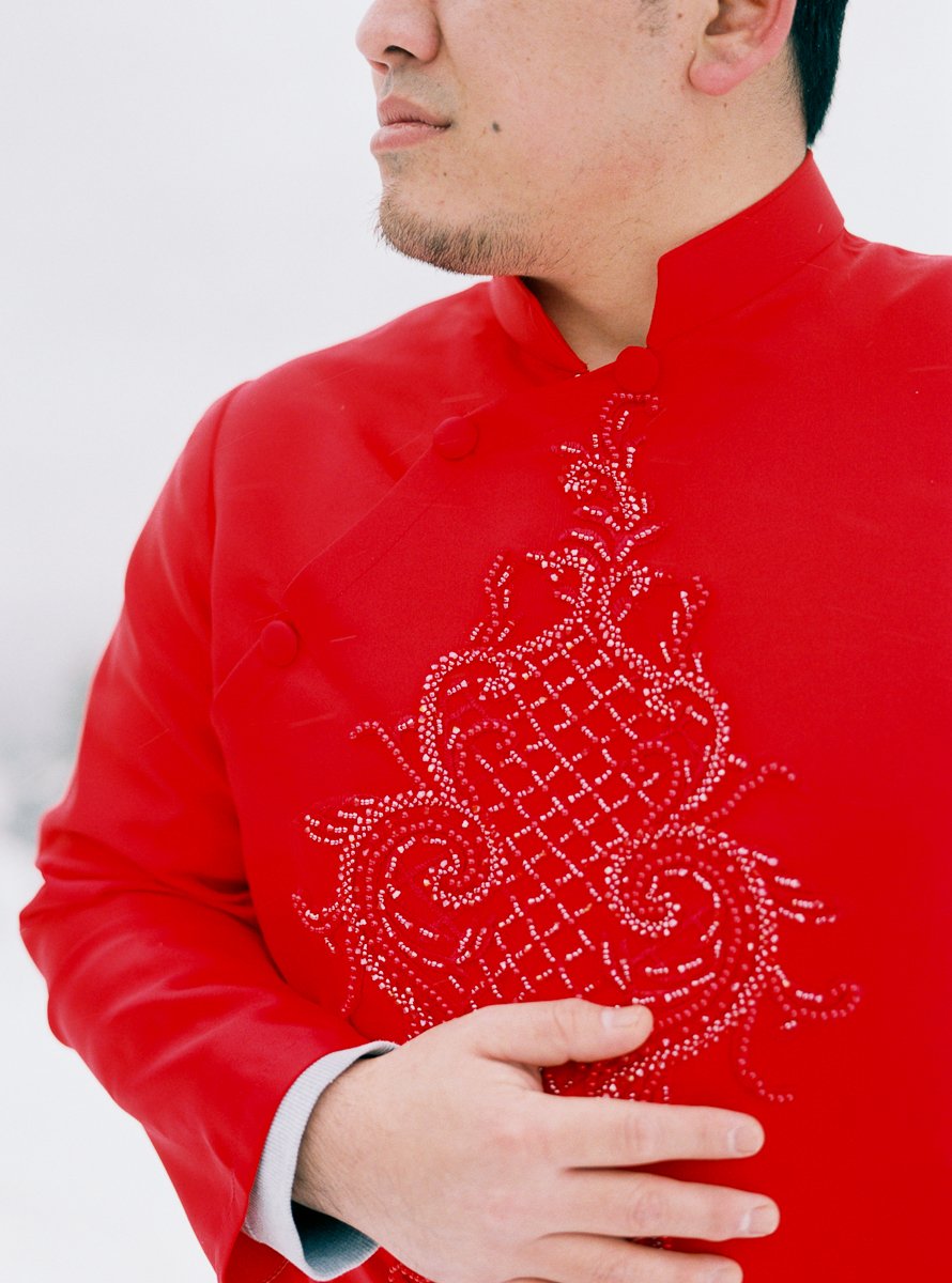 Close-up detail of groom’s red embroidered Vietnamese áo dài with hand resting on chest.