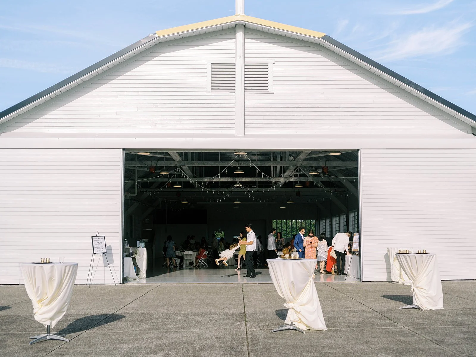 Exterior wide shot of the Pearson Air Museum hangar wedding venue with draped cocktail tables on the tarmac and guests mingling inside under string lights
