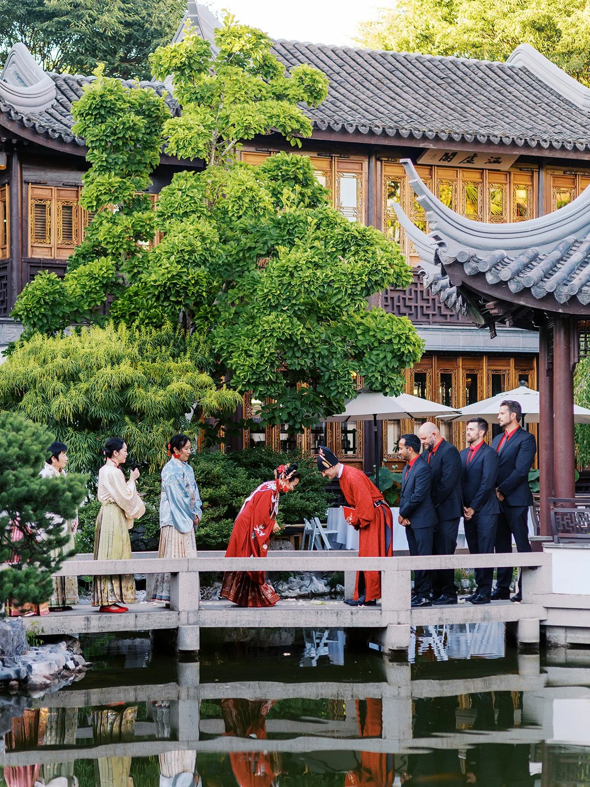 A wider view of the same bowing ceremony moment, the couple deeply bowed on the bridge with the full pavilion and sweeping roofline reflected in the calm water.