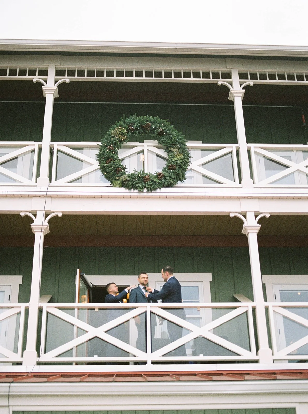 Groom stands on a balcony while two groomsmen help him get ready beneath a large evergreen wreath.