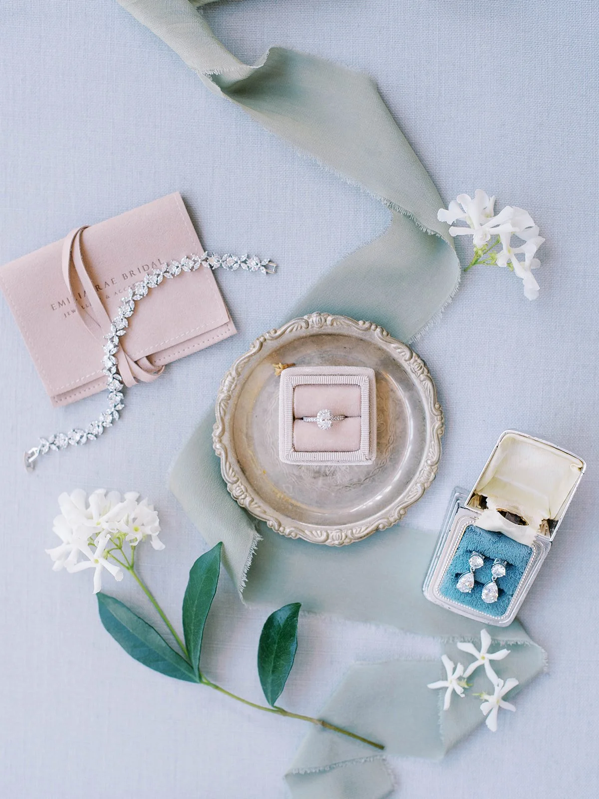Wedding jewelry flat lay with diamond ring, bracelet, earrings, silk ribbon, and white florals on soft blue linen at Abernathy Center