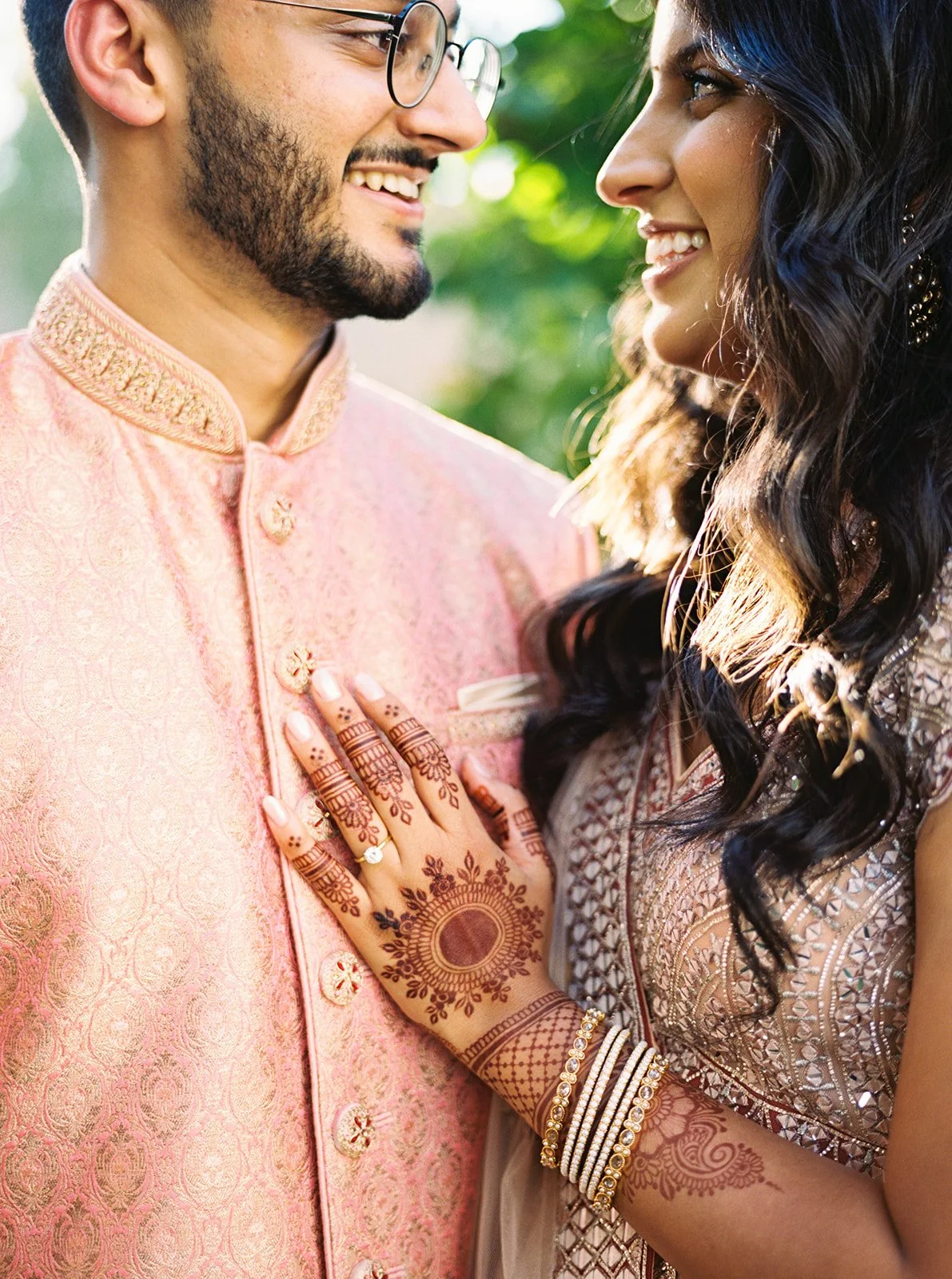 Bride’s mehndi-adorned hand resting on groom’s peach sherwani as they smile at each other.
