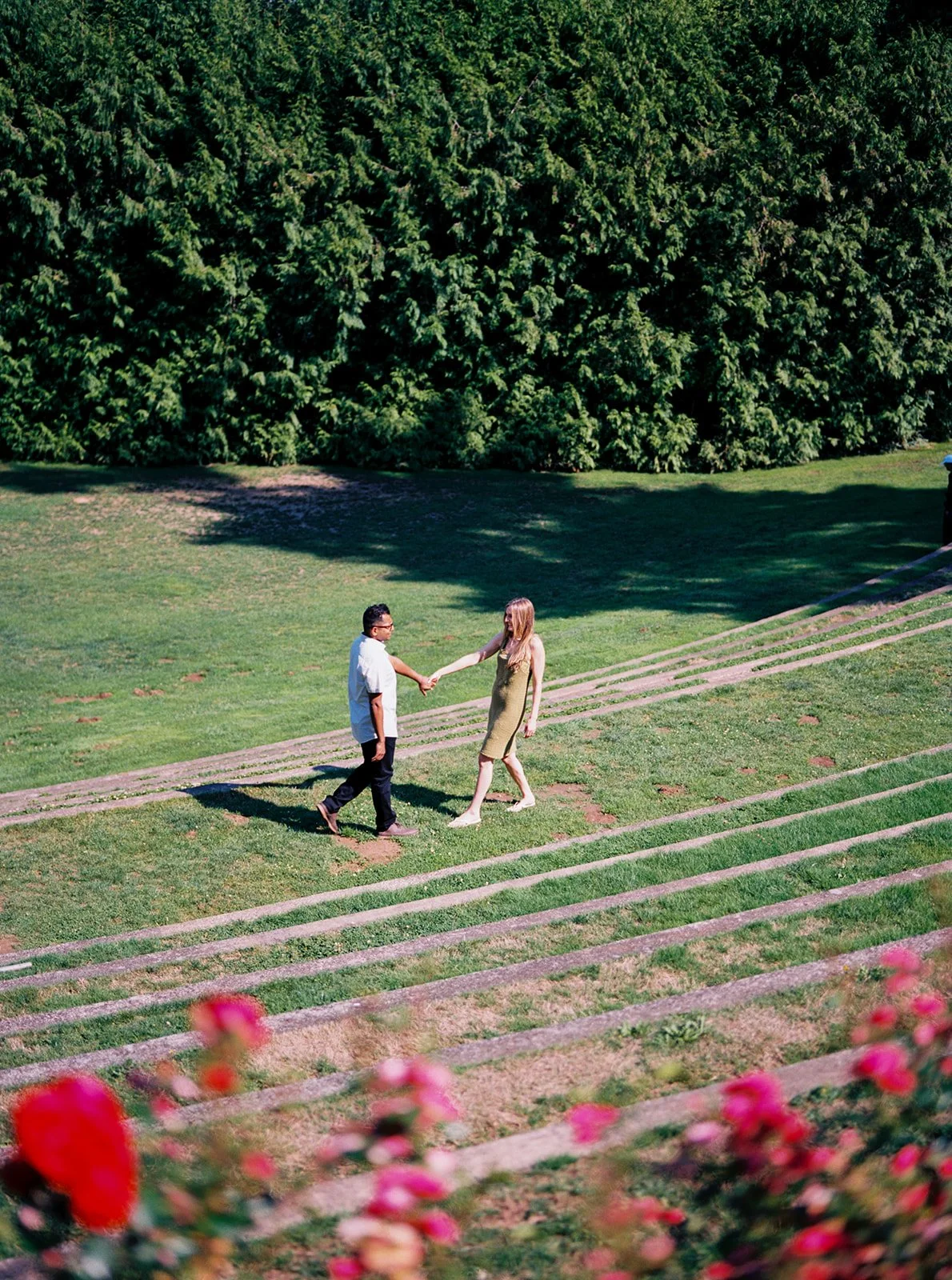 An aerial shot of a couple holding hands while walking across a terraced lawn, pink roses blurred in the foreground.