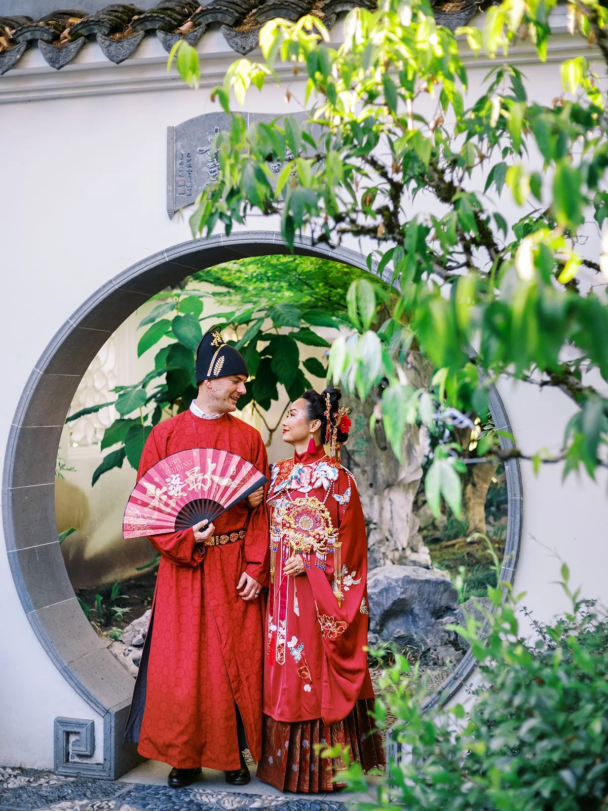 Bride and groom in red traditional Chinese wedding attire standing together inside a stone moon gate, surrounded by lush greenery at Lan Su Chinese Garden