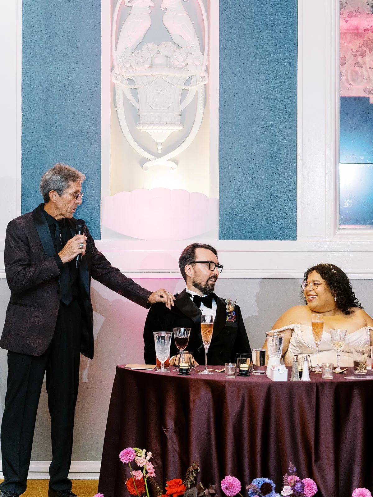 Guest giving a speech beside a seated couple at a decorated head table with burgundy linens and vibrant florals.