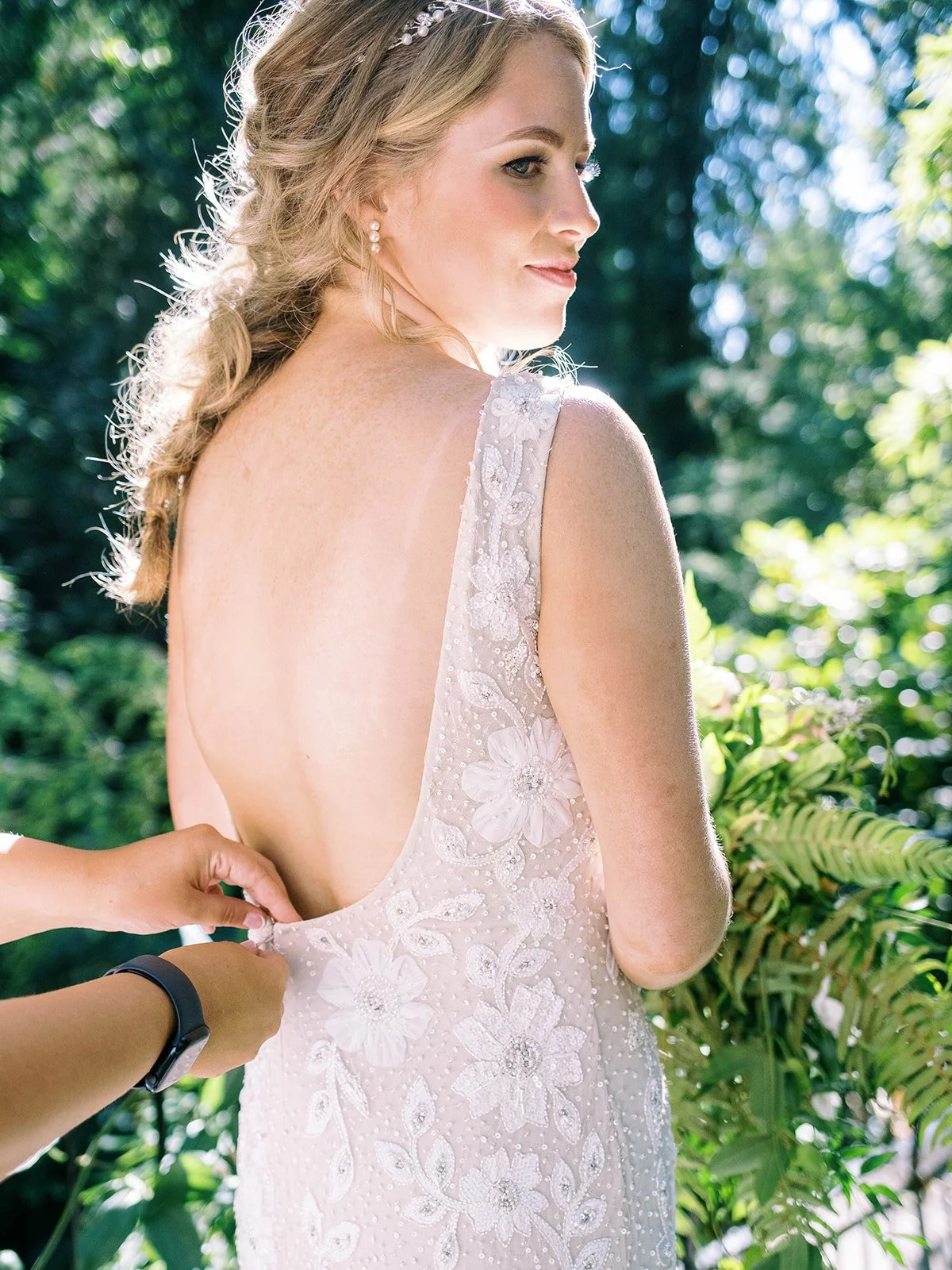 Bride in a backless floral wedding gown as someone fastens the dress outdoors.