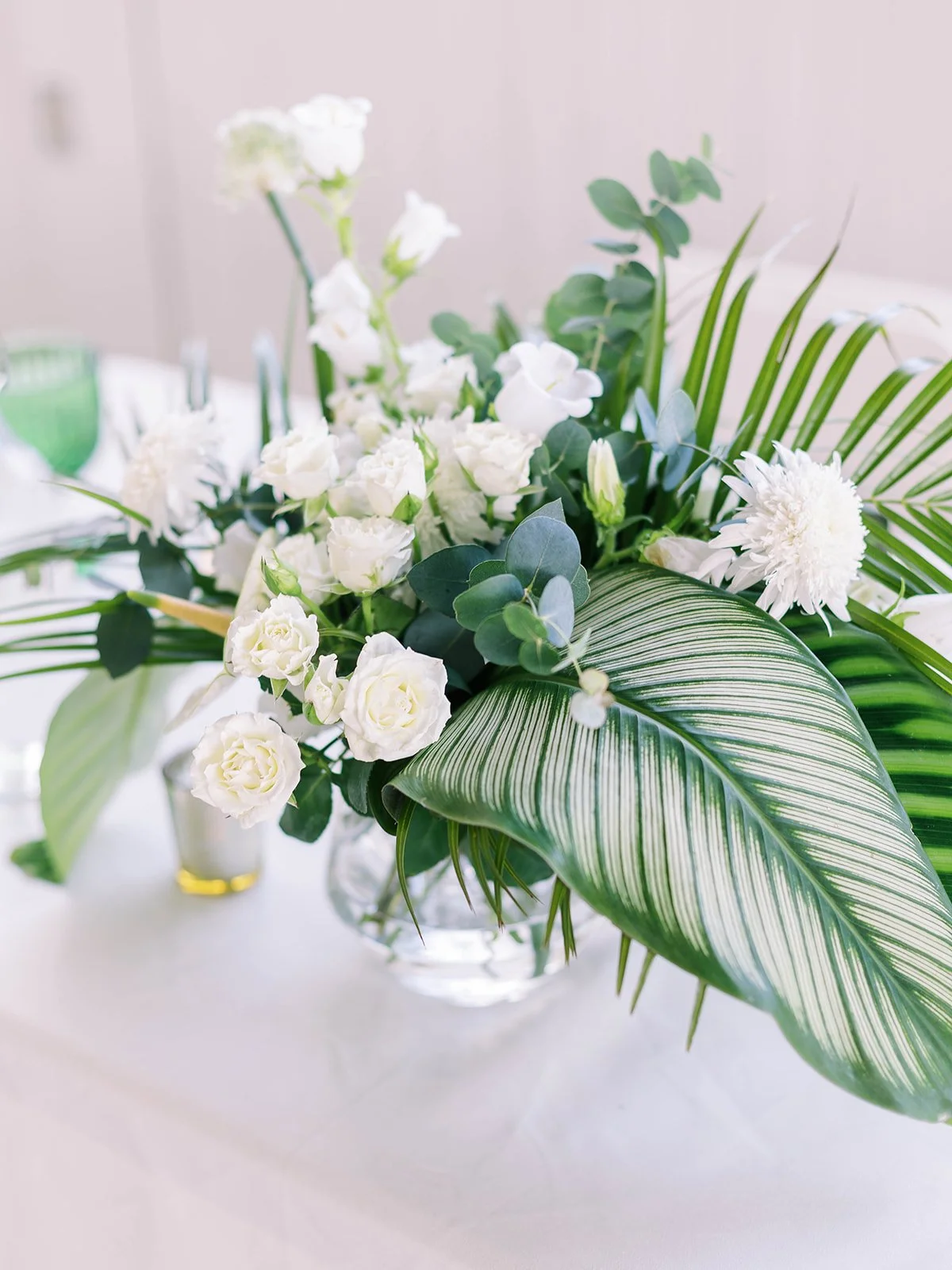 Close-up of a lush tropical wedding floral centerpiece with white roses, lisianthus, eucalyptus, and striped calathea leaves in a glass vase
