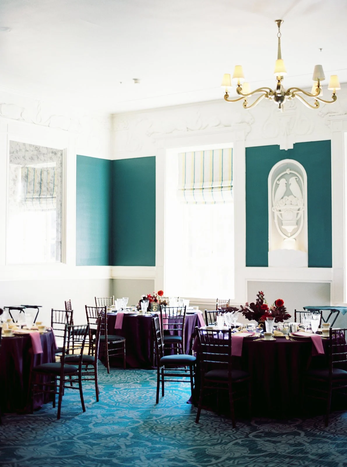 Elegant reception space with teal walls, chandeliers, and round tables set with burgundy linens and floral centerpieces.